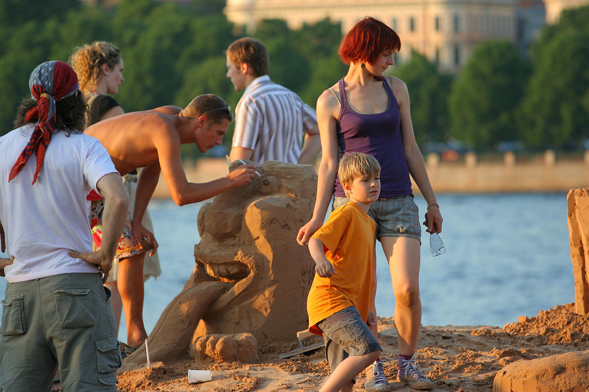 A group of people are building sand sculptures by a body of water on a sunny day, celebrating Oligarhu kapusvētki. One shirtless man is sculpting a figure, while a woman with red hair in a purple tank top and shorts stands near a young boy in an orange shirt. Others converse in the background.