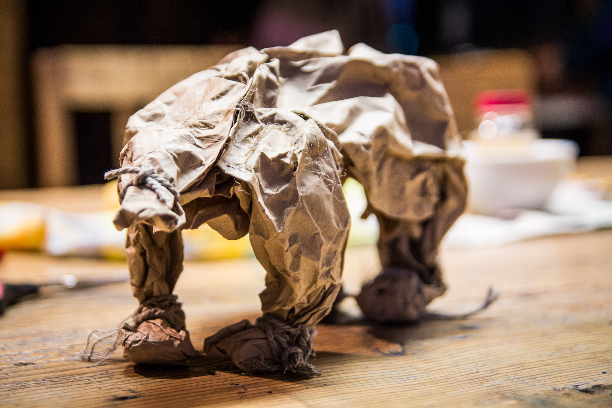 A close-up photograph displays a sculpture of an elephant made from crumpled brown paper. The elephant stands on a wooden table, with blurred background elements including a white bowl and red container. The texture of the paper and the detailed folds mimic the contours of an elephant's body, reminiscent of creations seen at the Papīra objektu festivāls.
