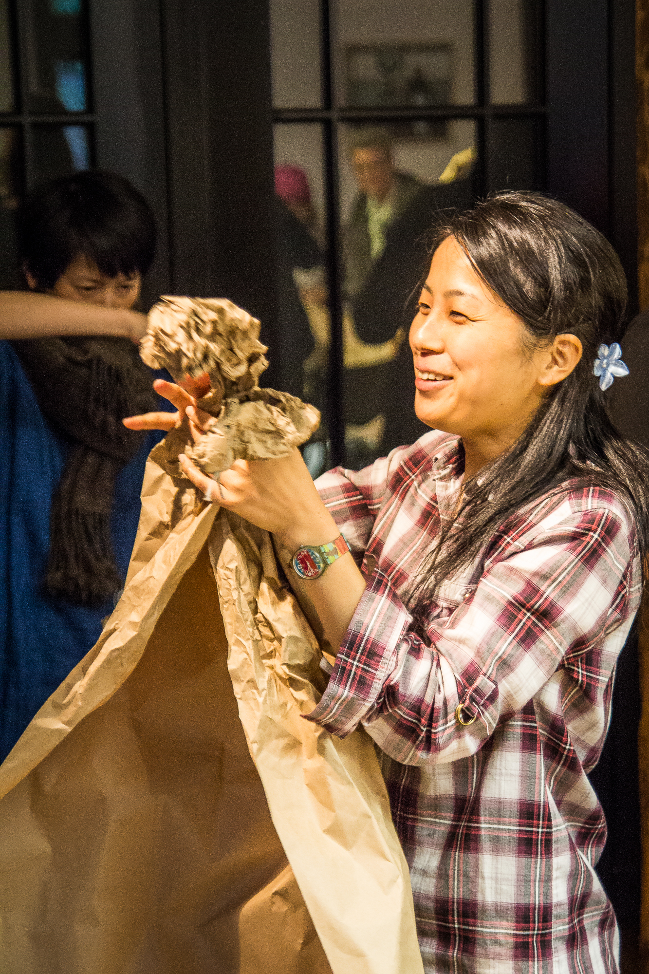 A woman with long hair, wearing a plaid shirt and a colorful wristwatch, joyfully crumples brown packing paper at the Papīra objektu festivāls. Behind her, another person with short hair in a blue shirt and dark scarf watches, covering their mouth with their arm. The background features a window reflecting figures in a room.