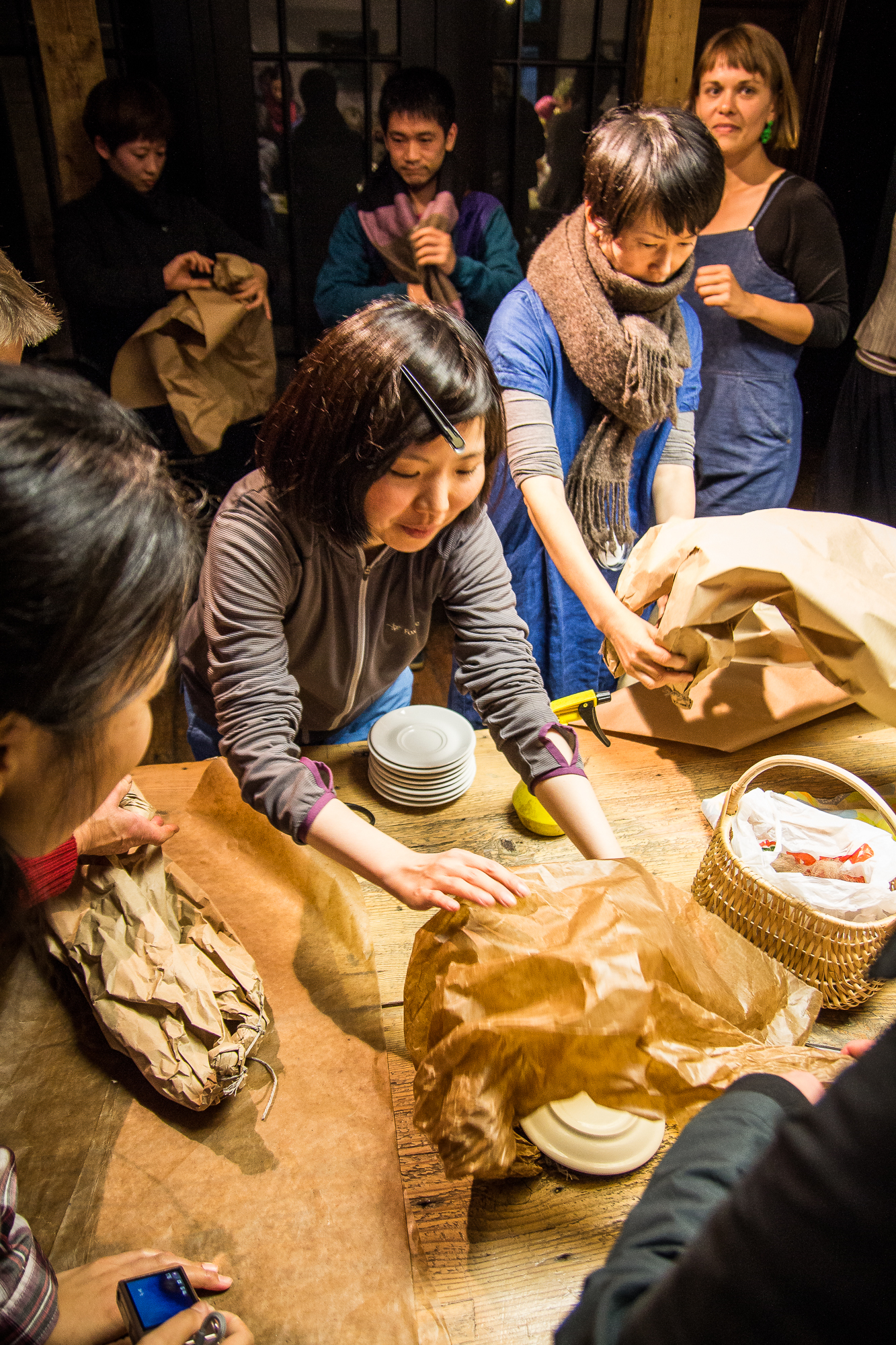 A group of people are gathered around a wooden table, unwrapping and organizing ceramic plates from brown paper packages. One person in the center, wearing a gray jacket, is reaching for a stack of plates. Others are assisting and observing. The atmosphere resembles a festivāls of Papīra objekti, collaborative and focused.