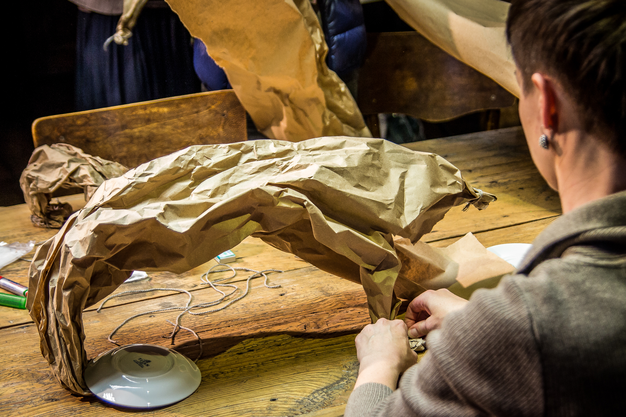 A person with short hair works on creating a sculpture using crumpled brown paper and a lamp base on a worn wooden table, as part of the Papīra objektu festivāls. Various craft materials and another partially seen individual are in the background, making for a lively and creative indoor setting.