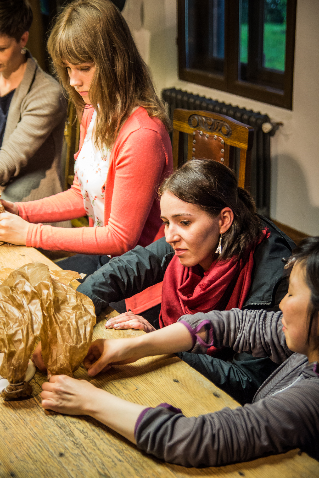 Four people are seated around a wooden table engaged in crafting papīra objekti. One woman in the foreground wears a red scarf and closely examines the paper. Another, in a pink cardigan, focuses intently on her work. The room has a cozy, rustic feel with a window and radiator in the background.