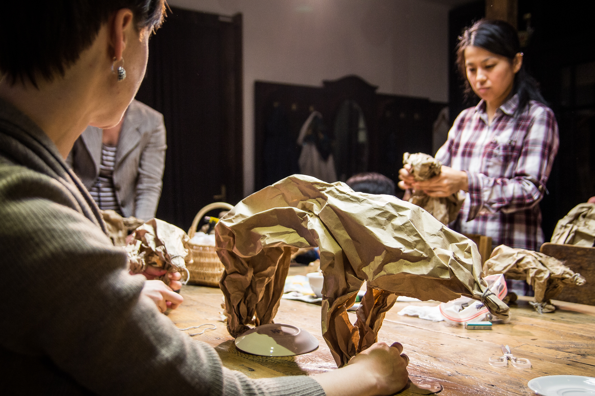 People crafting with brown paper at a wooden table. In the foreground, a person is shaping the paper into a structure. In the background, another individual in a plaid shirt is also working with the paper. The setting appears to be indoors with dim lighting and various crafting supplies are scattered—Papīra objektu festivāls in full swing.