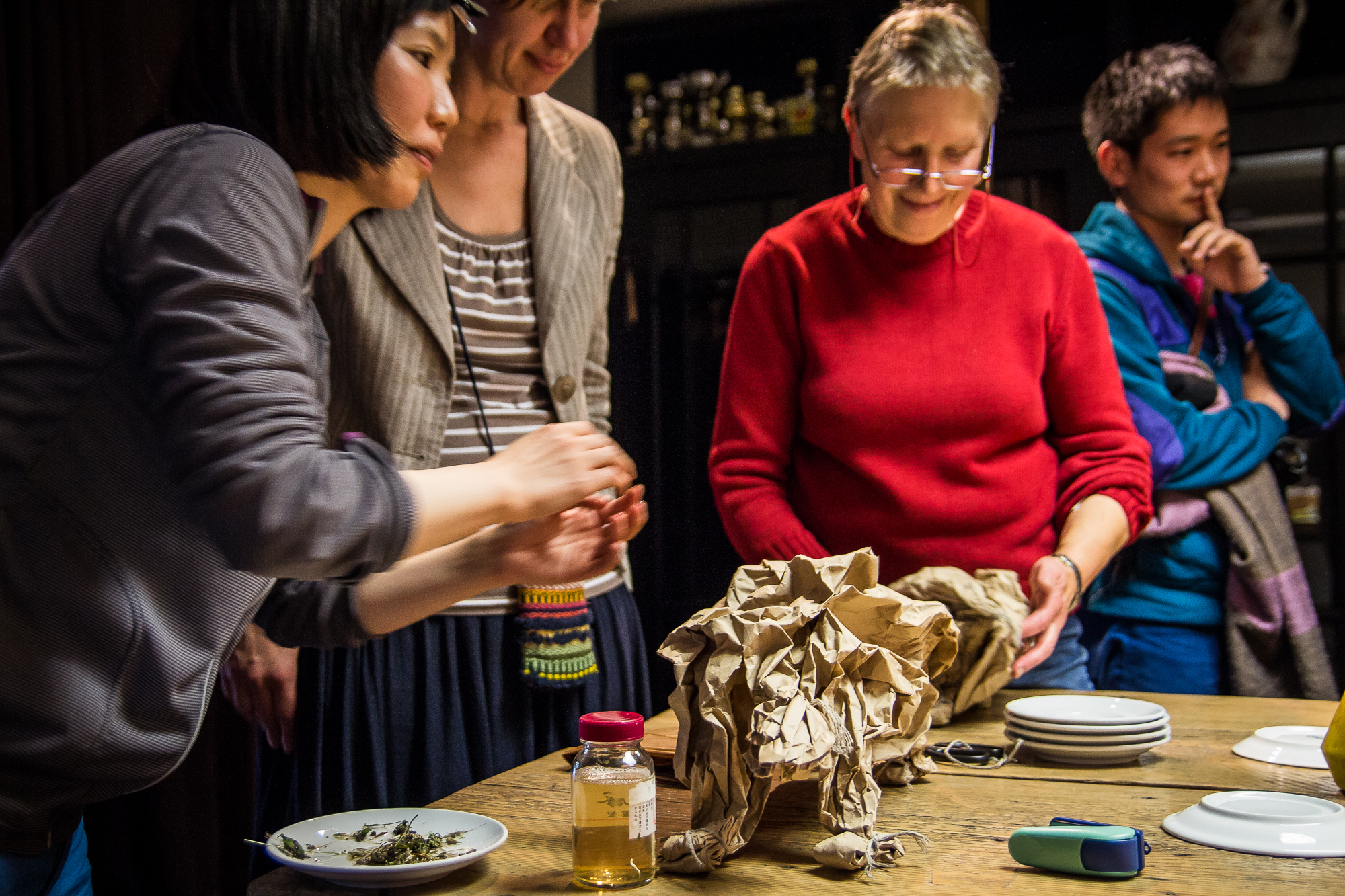 A group of four people stands around a wooden table. One person on the left is handling items wrapped in crumpled brūnais Papīra. Various objektu, including plates, a jar with yellow liquid, a canister, and dried leaves, are spread out. The mood appears to be focused and collaborative as if preparing for a festivāls.