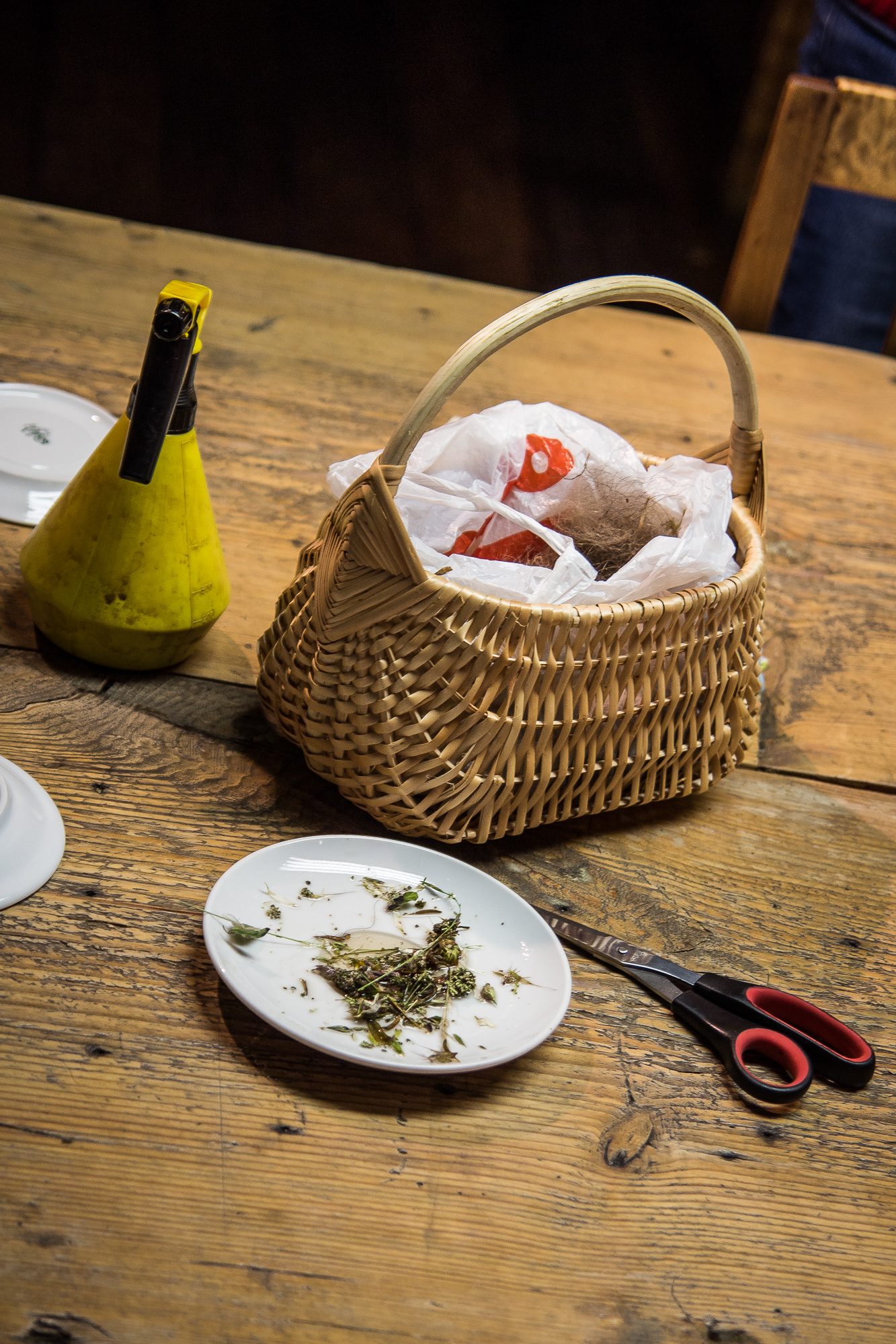 A wicker basket, partially filled with wrapped items, rests on a wooden table. Beside it, there is a yellow oil dispenser. In the foreground, a white plate holds herbs, and a pair of red-handled scissors lies nearby. A flyer for the Papīra objektu festivāls peeks out from behind the basket.