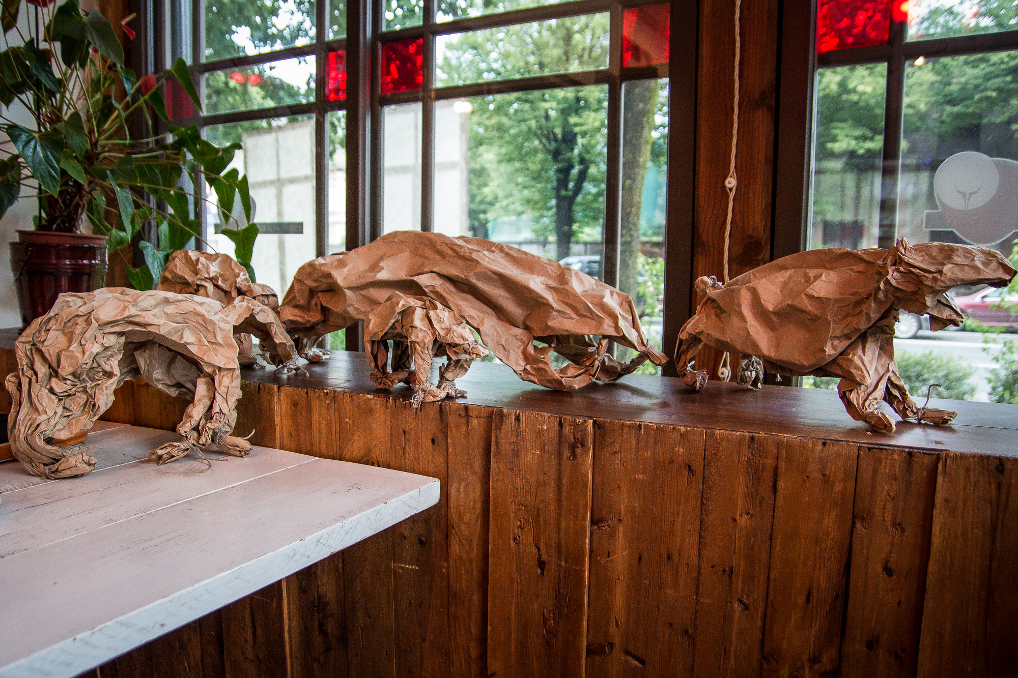 Four animal sculptures crafted from crumpled brown paper bags are displayed on a wooden windowsill. The animals resemble a bear, a dog, and other quadrupeds. Green potted plants and large windows with views of trees and buildings are visible in the background, evoking a Papīra objektu festivāls vibe.