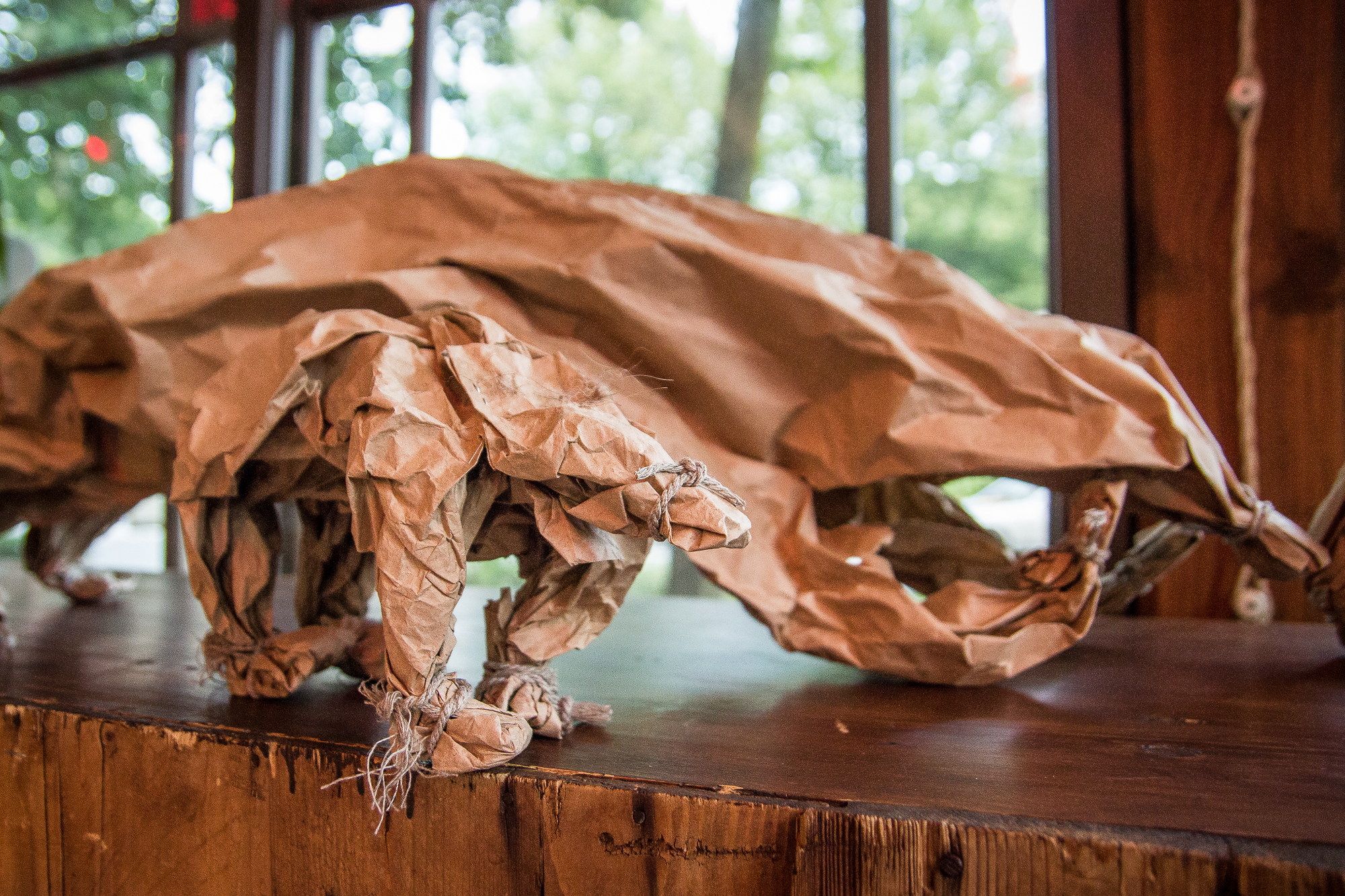 A sculpture of a bear made from crumpled brown paper is displayed on a wooden surface. The detailed paper folds create a lifelike representation of the bear, positioned against a background of large windows showcasing a green, leafy outdoor scene, evoking the essence of the Papīra objektu festivāls.