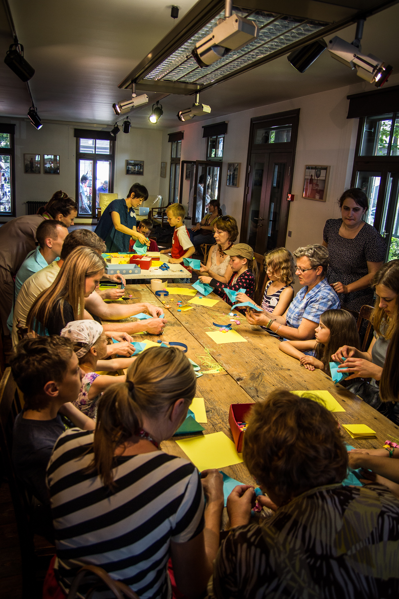 A group of adults and children gather around a long wooden table in a bright, well-lit room. Engaging in a craft activity for the festivāls, they are folding colorful papīra and creating art. The participants appear focused and engaged. The room has large windows, framed pictures, and light fixtures on the ceiling.