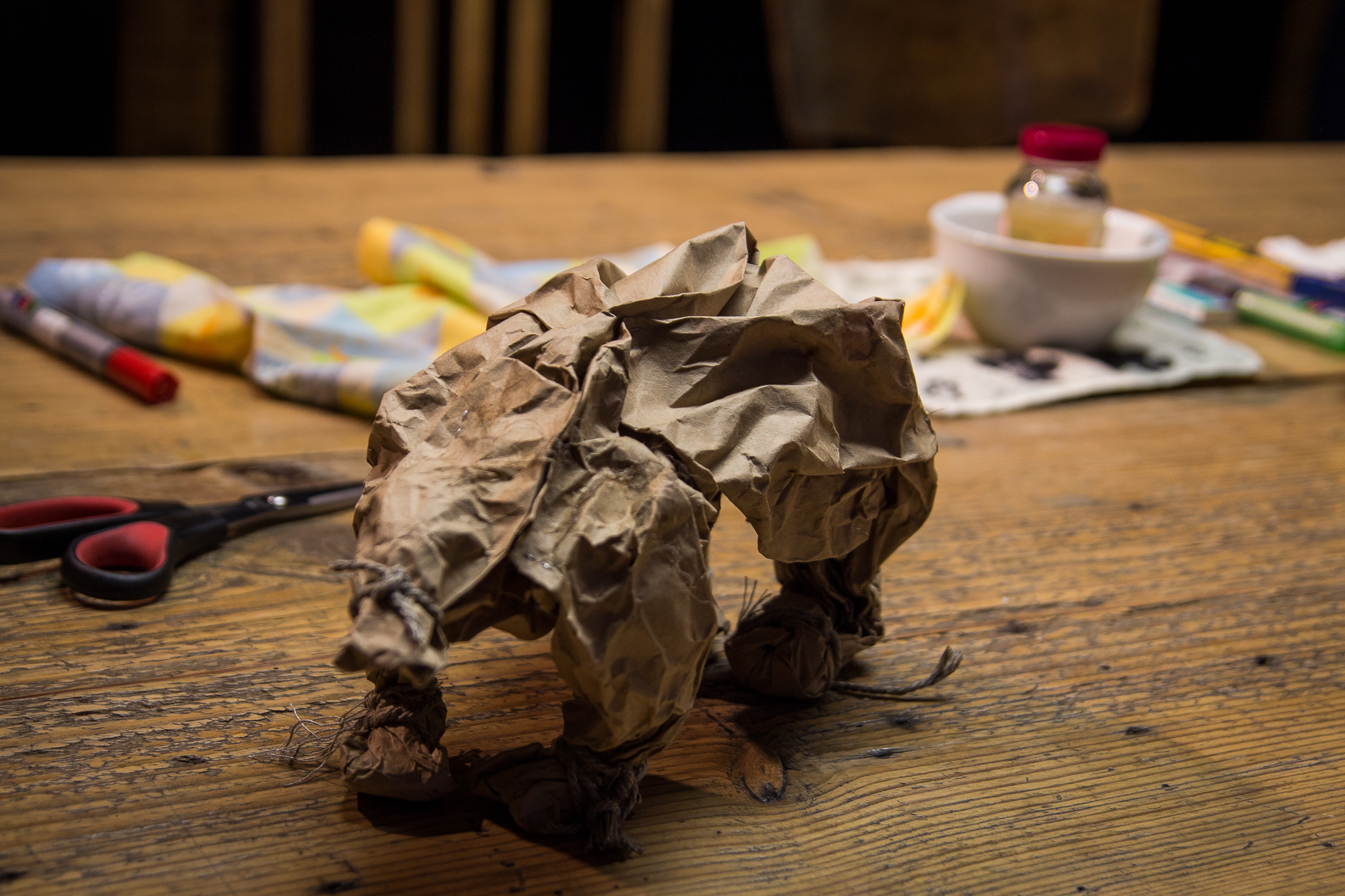 A crumpled paper sculpture of a bear stands on a wooden table. To the left, a pair of red-handled scissors lies open. In the background, various art supplies, including pencils, cloth, and a small jar, are scattered around, reminiscent of an art festivāls with wooden chairs visible further back.
