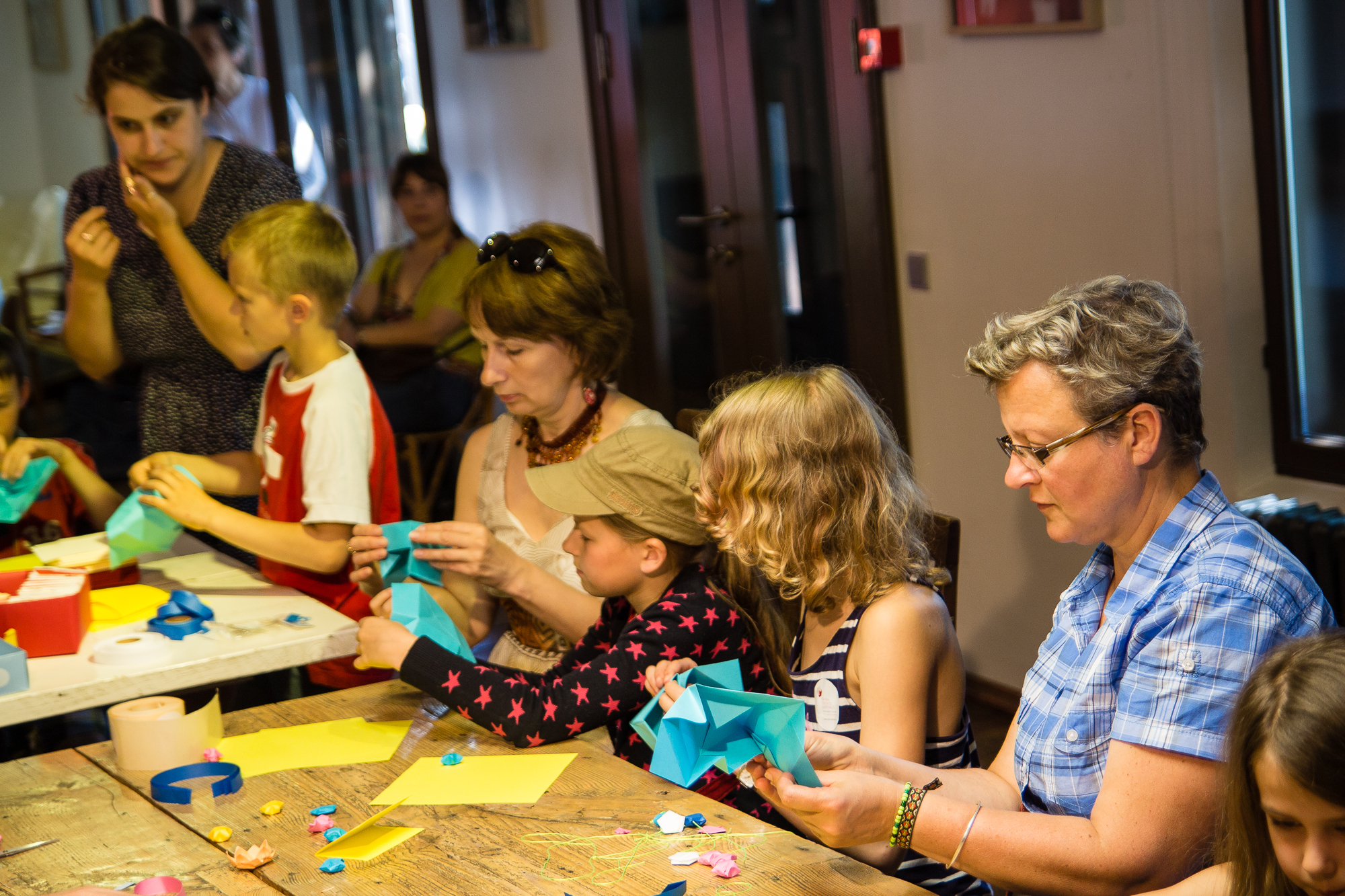 A group of children and adults are sitting around a wooden table at the festivāls, engaging in craft activities. They are folding colorful origami papers, transforming them into intricate papīra objekti. The table is scattered with paper pieces, tape, and craft tools. In the background, other people are observing. The scene is lively and creative.