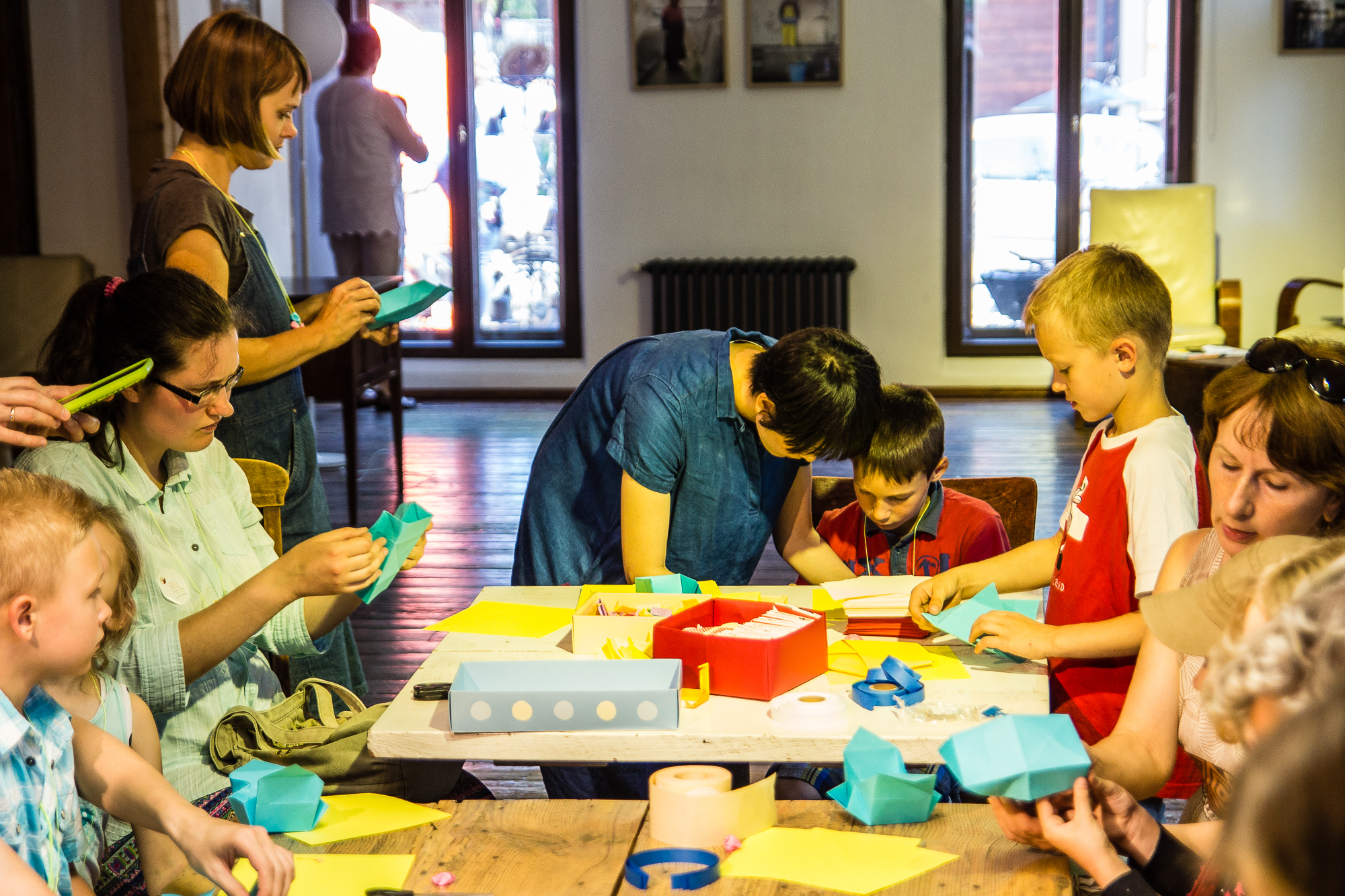 A group of children and adults are gathered around a table at the Papīra Objektu Festivāls, engaged in arts and crafts. Some are folding colored paper, while others assist the children. The room is well-lit with large windows in the background, allowing natural light to fill the space. Various supplies are scattered on the table.