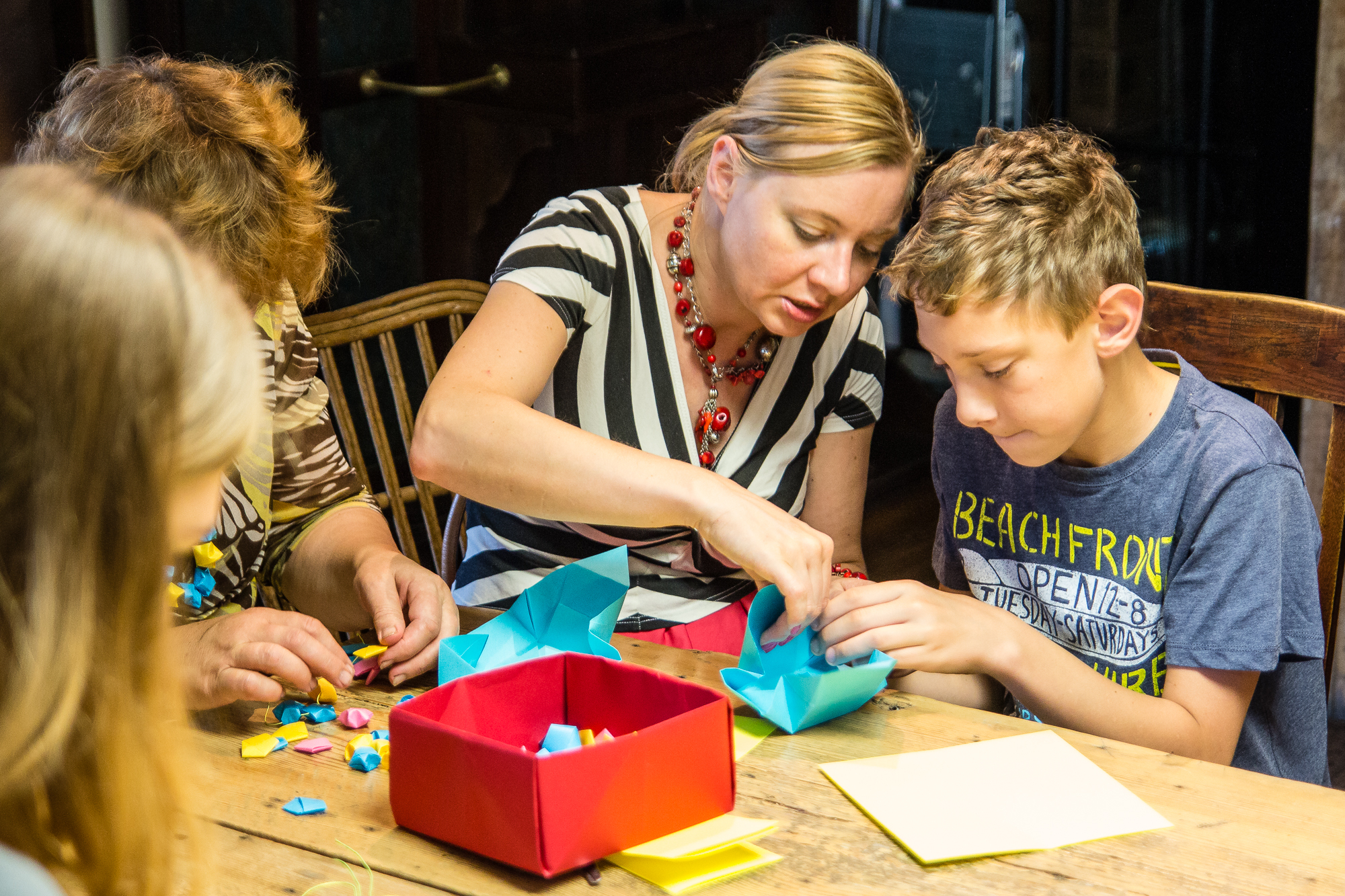 A woman in a striped shirt helps a boy in a blue t-shirt with an origami project during a papīra objektu festivāls. They are seated at a wooden table cluttered with colorful paper, and a red box is in front of them. Two other people, partially visible, are also engaged in folding paper on the left side of the image.