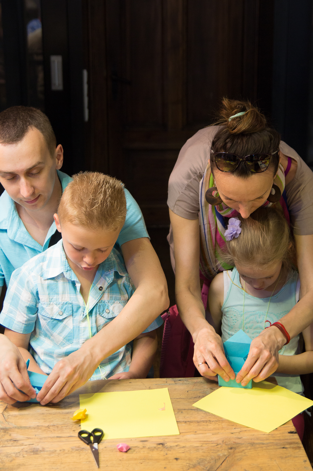 A man and a woman assist two young children with an art project at a wooden table. They are folding and cutting colorful paper, perhaps preparing for the upcoming Papīra objektu festivāls. Scissors and other craft materials are scattered on the table. The adults are focused on helping the children, who appear engaged in the activity.