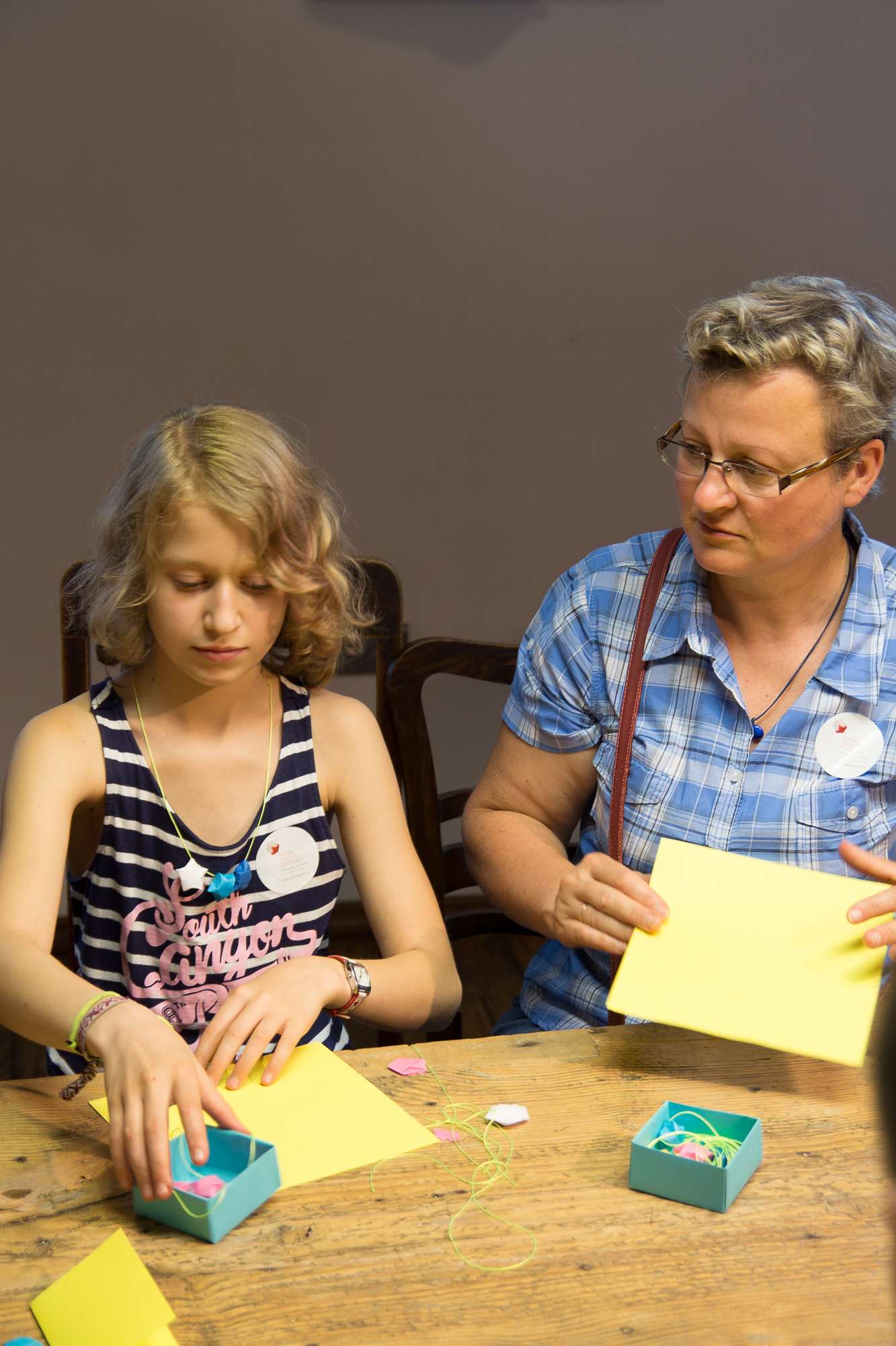 A young girl and an older woman are sitting at a wooden table engaged in a crafting activity, preparing for the upcoming festivāls. The girl, dressed in a striped tank top, is focused on her papīra objektu with small items, while the woman in blue plaid supervises, holding a yellow piece of paper.