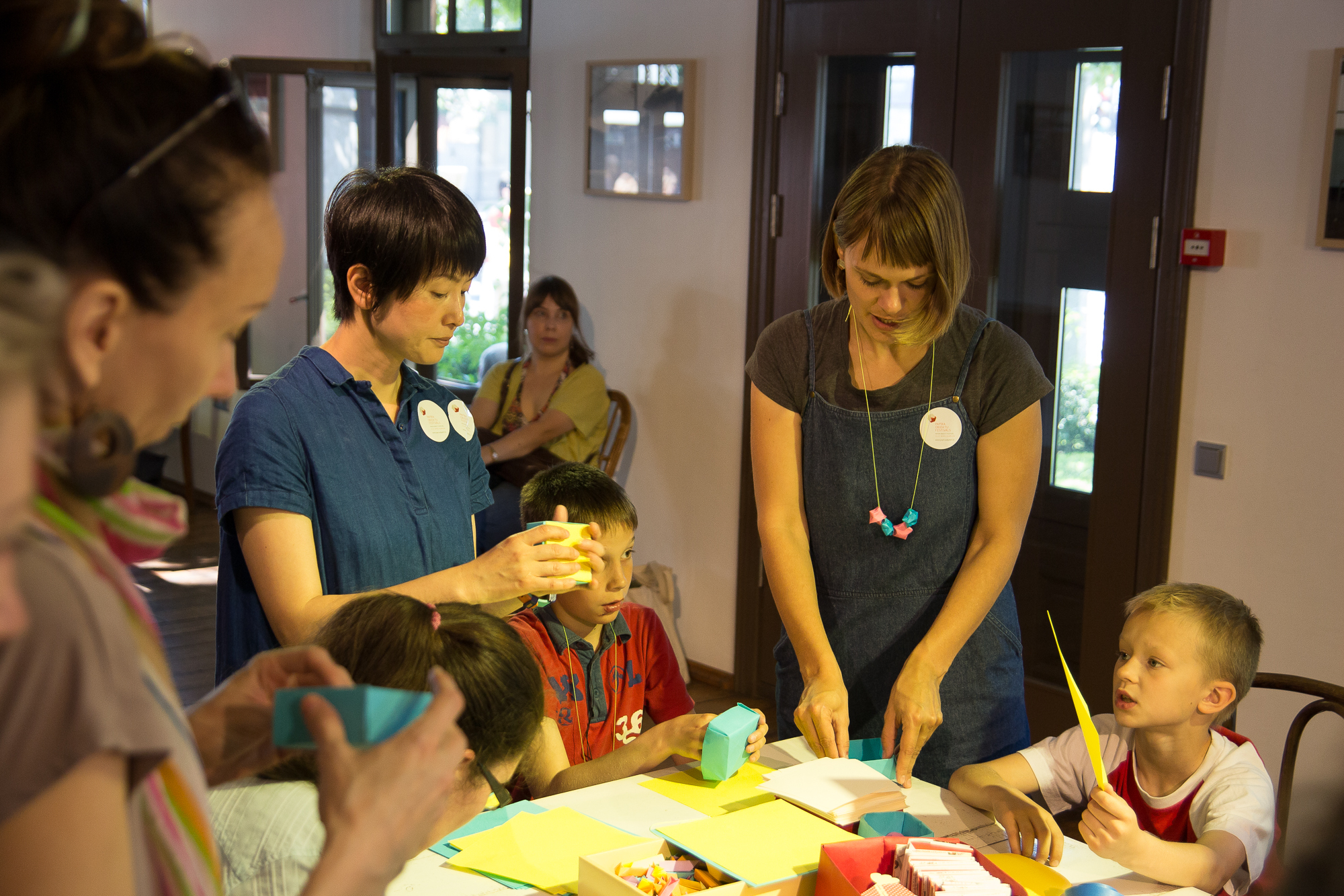 A group of children and adults is engaged in a crafts workshop inside a well-lit room with wooden furniture. Two adults assist the children, who are focused on colorful Papīra and craft materials on a large table. Various completed objektu projects are visible, and everyone looks absorbed in their activity.