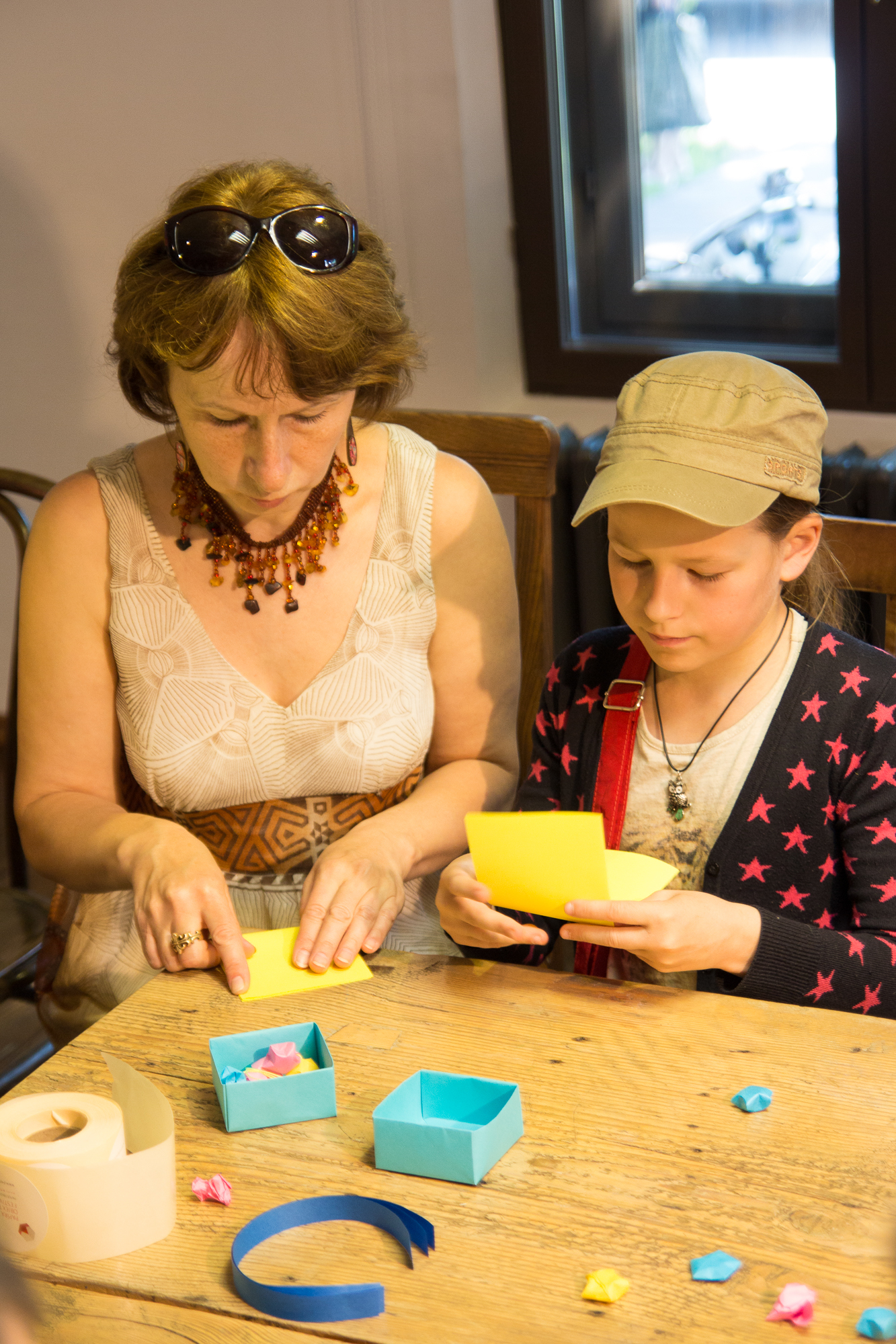 A woman and a child are engrossed in crafting papīra objektu at a wooden table. The woman, wearing sunglasses on her head, a light dress, and a beaded necklace, guides the child in a star-patterned cardigan who mirrors her actions. On the table are colorful papers, small boxes, and crafting tools.