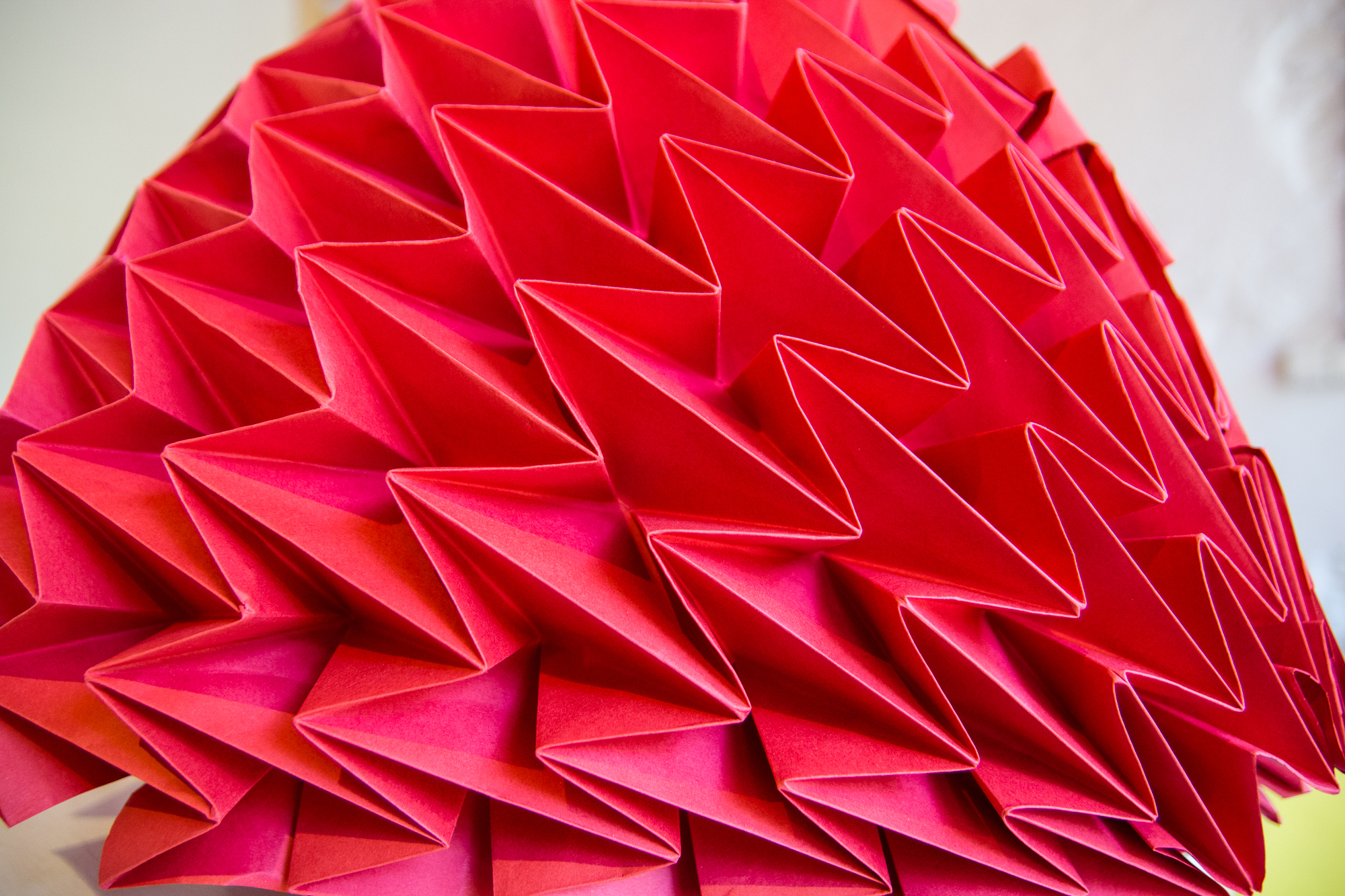 Close-up of an intricate red origami sculpture featured at the Papīra objektu festivāls, showcasing numerous sharp, angular folds and geometric patterns. The 3D paper artwork presents a complex, abstract form with layered triangular shapes creating a textured and vibrant surface.