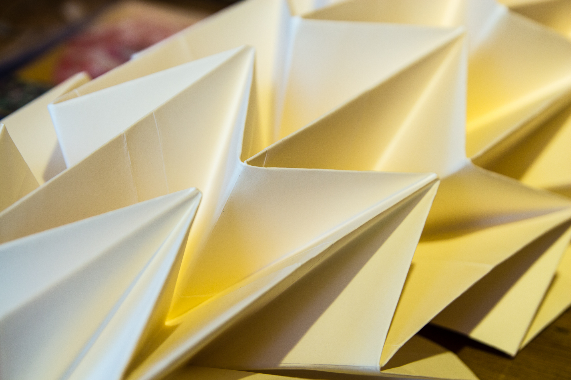 Close-up of intricately folded yellow Papīra origami creating a geometric, tessellated pattern. The folded shapes form sharp angles and interconnected points, casting soft shadows and creating a textured, three-dimensional appearance. The background is blurred and out of focus.