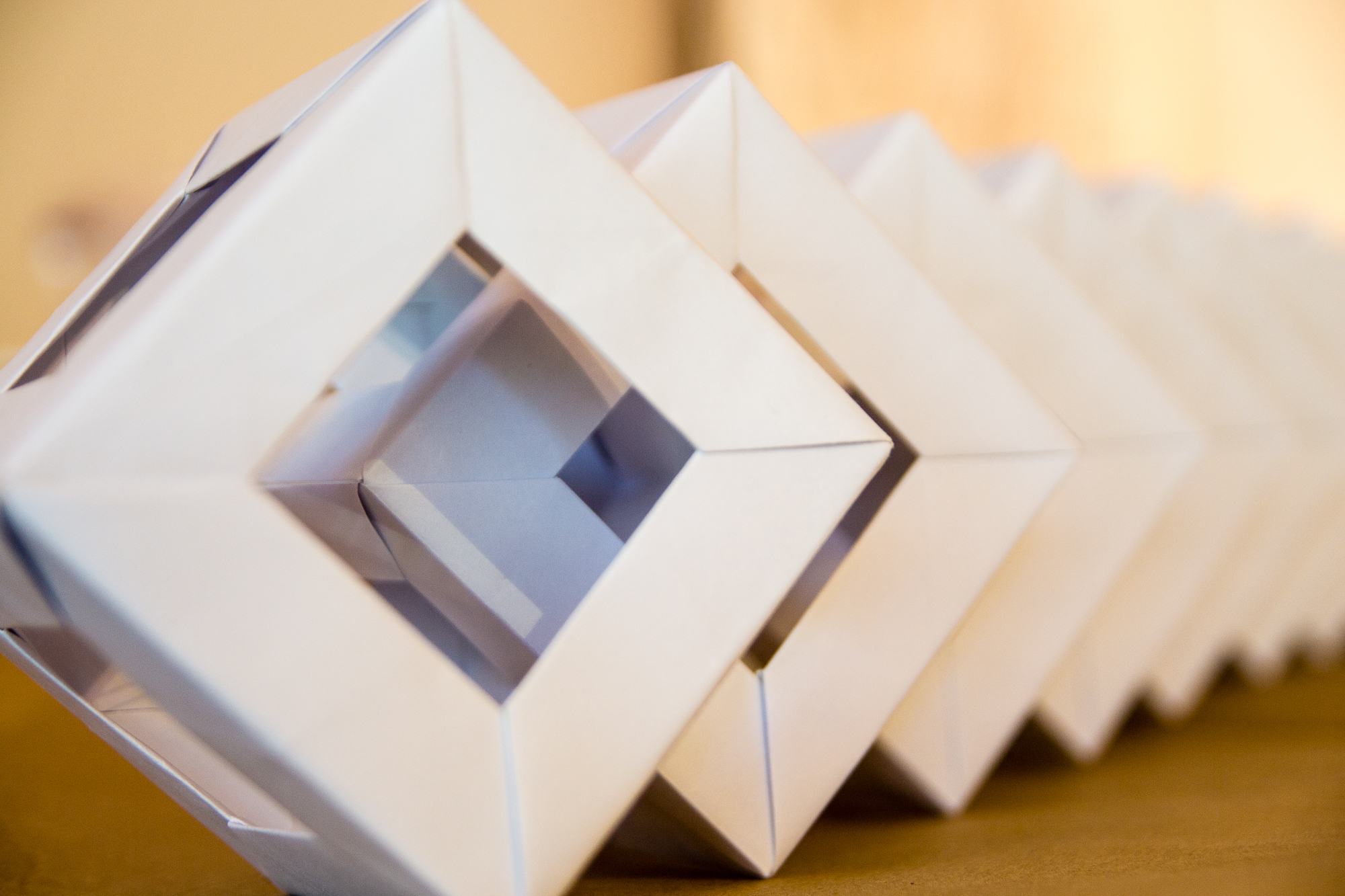 Close-up image of an abstract origami sculpture featuring a series of interconnected geometric shapes. Each shape consists of white paper folded into diamond-like frames, creating a repetitive, three-dimensional pattern. The background is blurred, focusing attention on the papīra objektu festivāls structure.