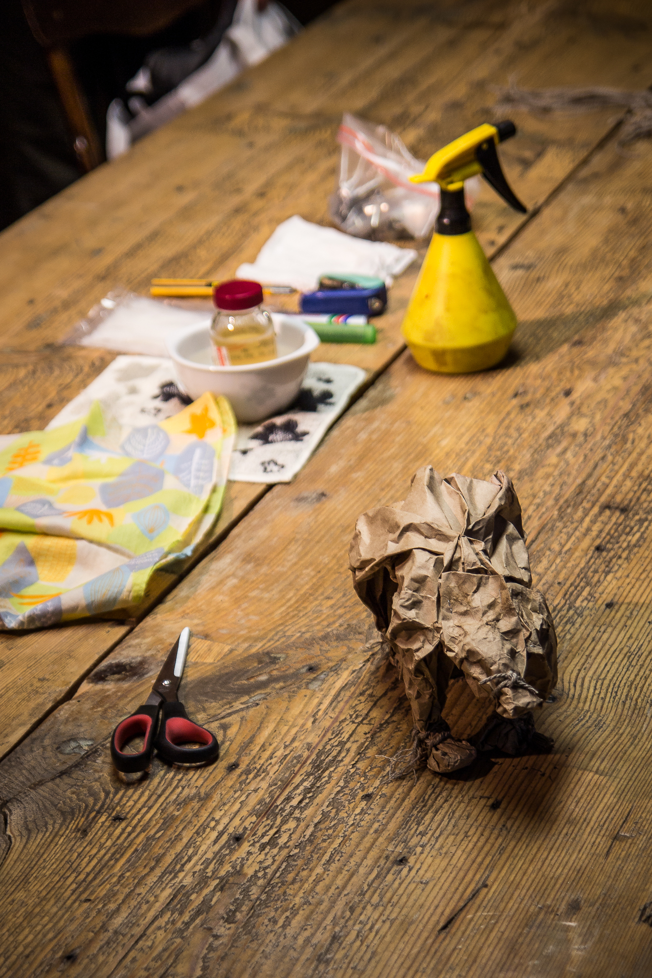 A wooden table with various items spread across: a pair of red and black scissors, a cloth with a fruit print, a crumpled brown Papīra bag, a yellow spray bottle, and a white bowl with small jars and plastic items next to a plastic bag. The background is softly lit, creating a warm ambience.