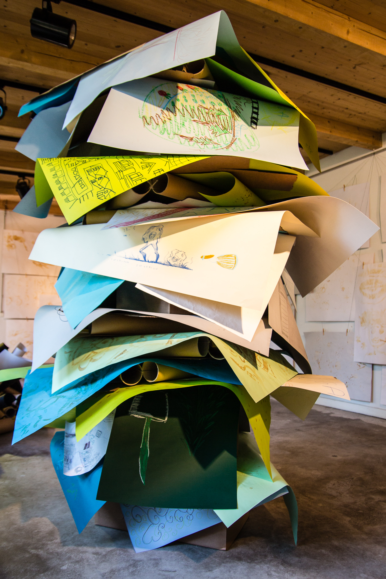 A large, colorful sculpture made of stacked, overlapping sheets of paper stands in an art gallery as part of the Papīra objektu festivāls. Each sheet features various doodles, drawings, and sketches in multiple colors. The artwork is displayed in a spacious room with wooden ceiling beams and unfinished walls adorned with additional drawings.