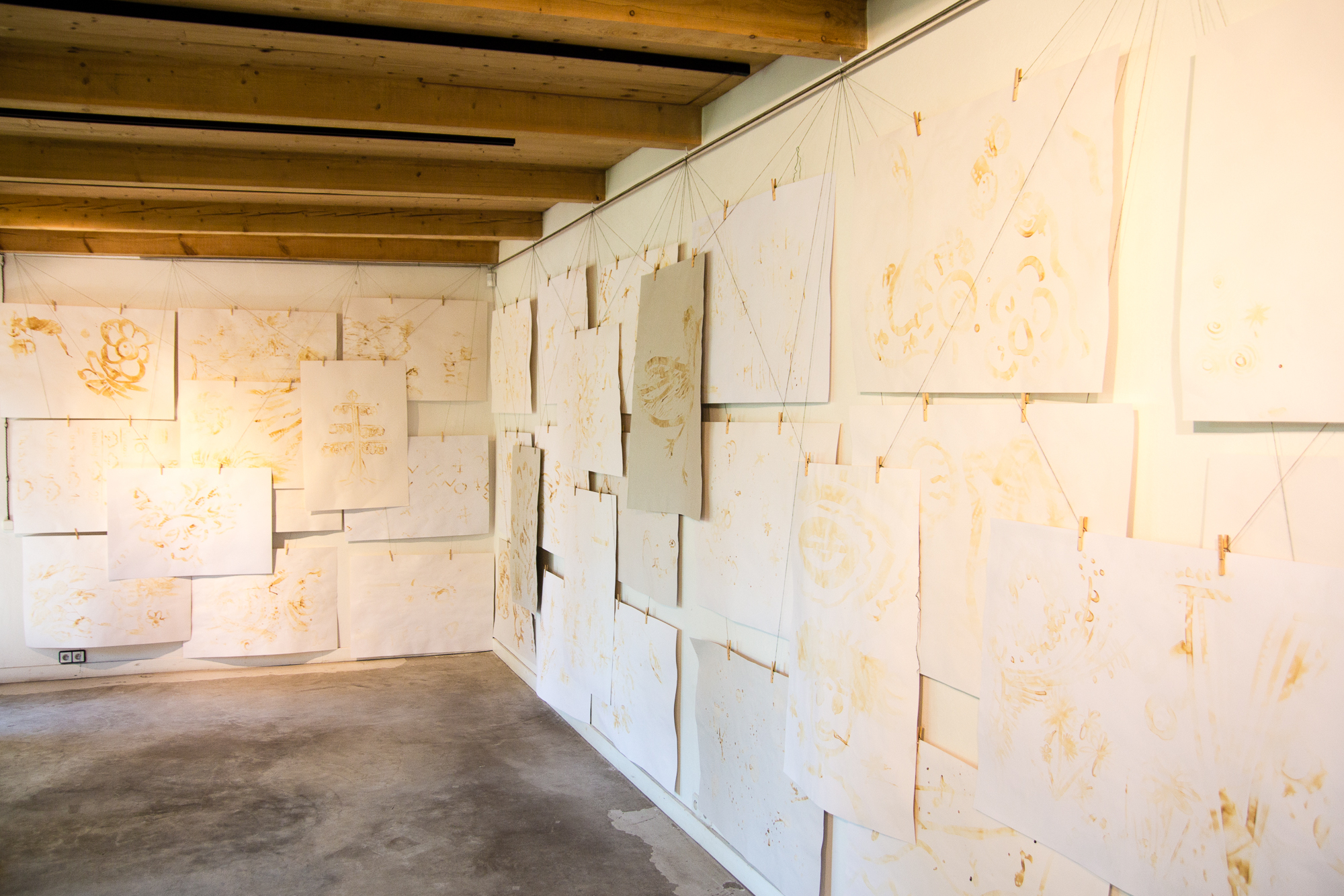 A gallery room with wooden ceiling beams displays numerous blank white canvases of various sizes, reminiscent of a Papīra objektu festival. The canvases are hung on white walls using clips and thin cords, creating a layered and overlapping effect. The concrete floor and lighting cast intriguing shadows on the canvases.