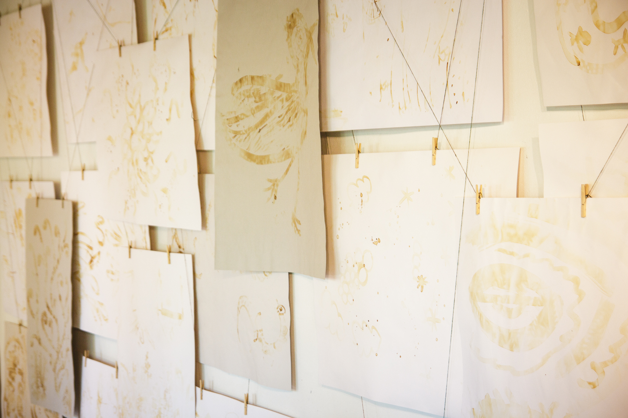A wall displays numerous sheets of paper with light brown sketches and abstract designs, resembling a Papīra objektu festivāls. The papers are pinned with clothespins to strings. Various loose, freeform drawings give a sense of creativity and experimentation. Light filters in, softly illuminating the artwork.