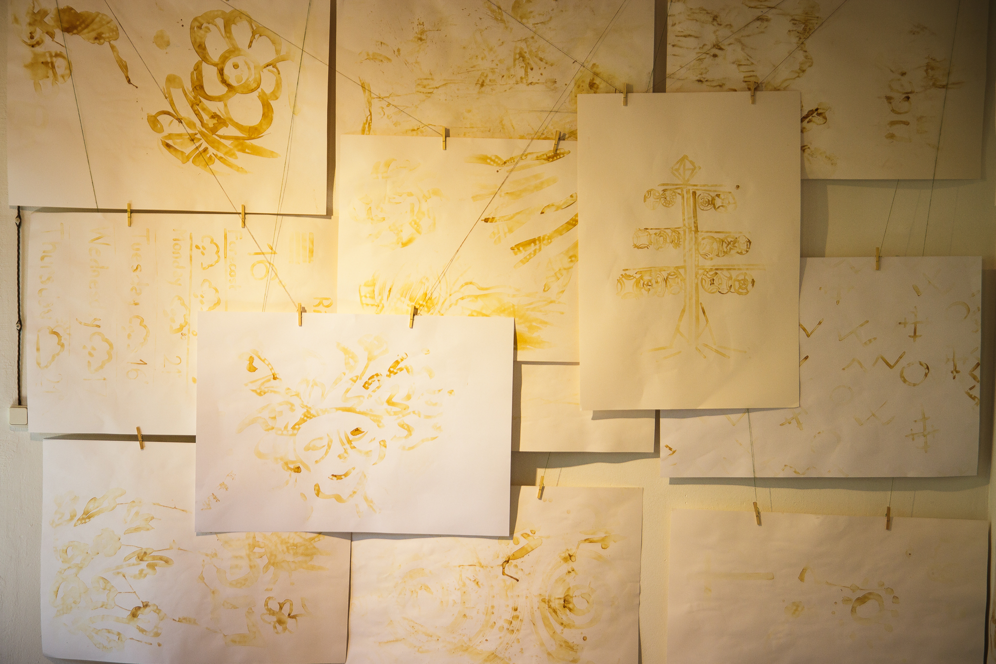 Several sheets of white papīra with various yellow/golden abstract drawings are displayed on a wall. The papers are pinned in an overlapping fashion, showcasing some floral, geometric, and symmetrical designs. The warm lighting enhances the golden hues of the artwork, creating a festivāls of visual delight.