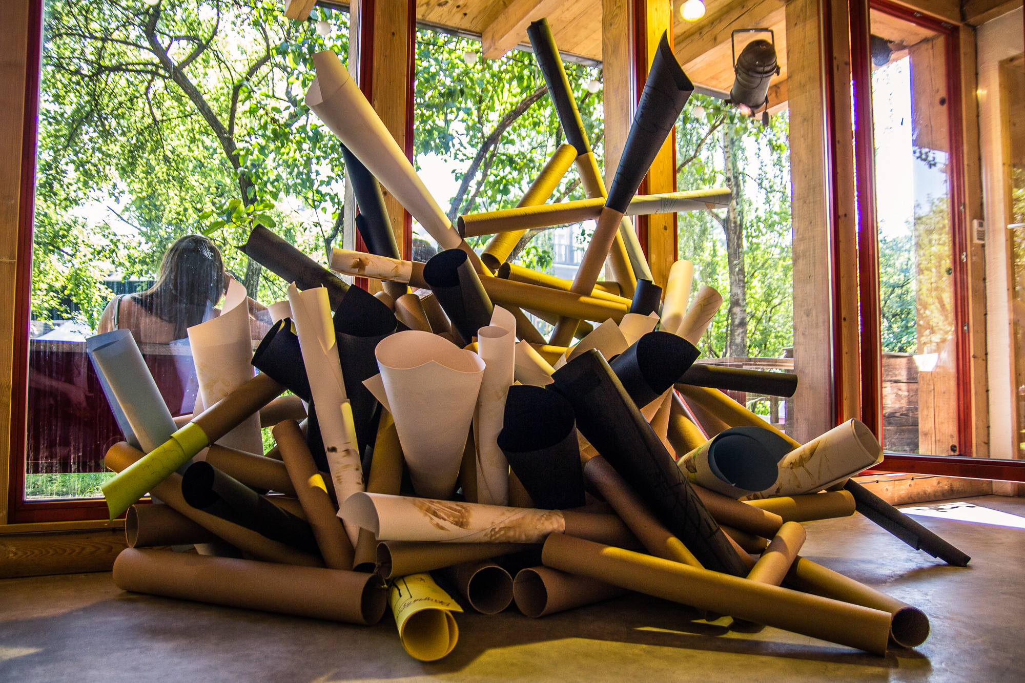 A large pile of cylindrical objects made of various materials and colors sits inside a room with floor-to-ceiling windows. Sunlight streams through the windows, illuminating the objects and casting shadows. Outside, green trees offer a vivid contrast to this creative indoor scene at the Papīra Objektu Festivāls.