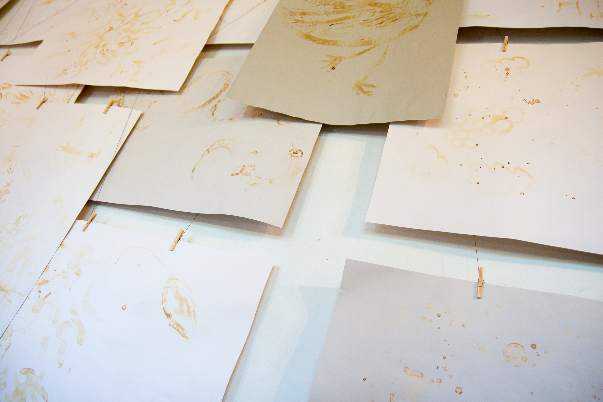 Several sheets of paper with various light brown ink sketches are pinned to a wall. The sketches include abstract and incomplete drawings, reminiscent of works from the Papīra objektu festivāls. The pages overlap slightly, creating a layered appearance, secured by small wooden clips attached to a string on the wall.
