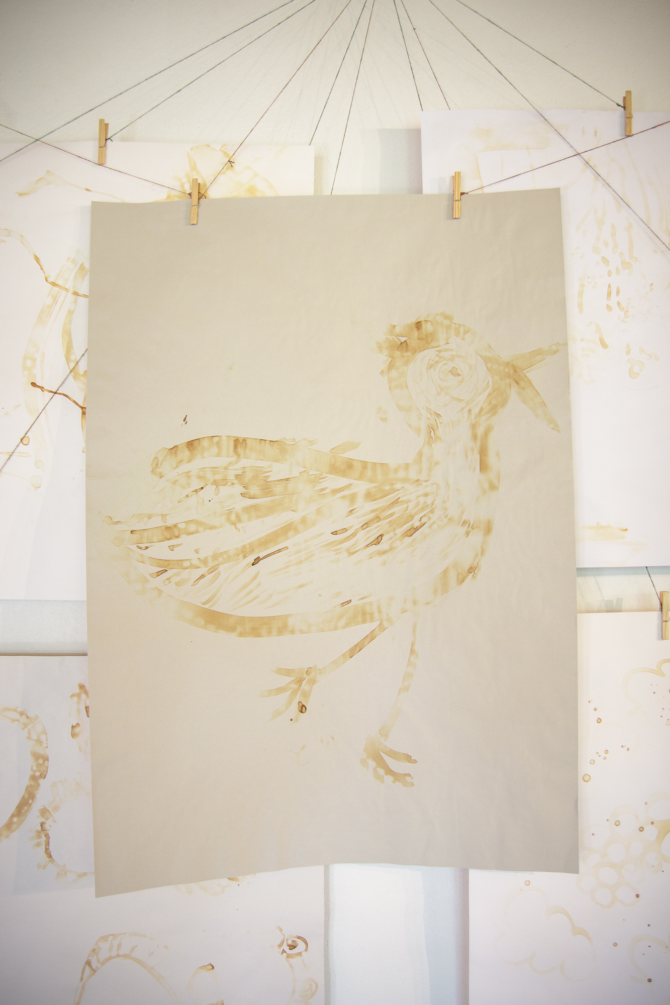 A drawing of a bird depicted using broad, flowing brushstrokes in sepia tones is displayed. The bird stands in profile on a large sheet of beige paper, which is secured to a line by two clothespins. Additional sheets with abstract designs hang around it like Papīra objektu festivāls, creating a gallery-like setting.