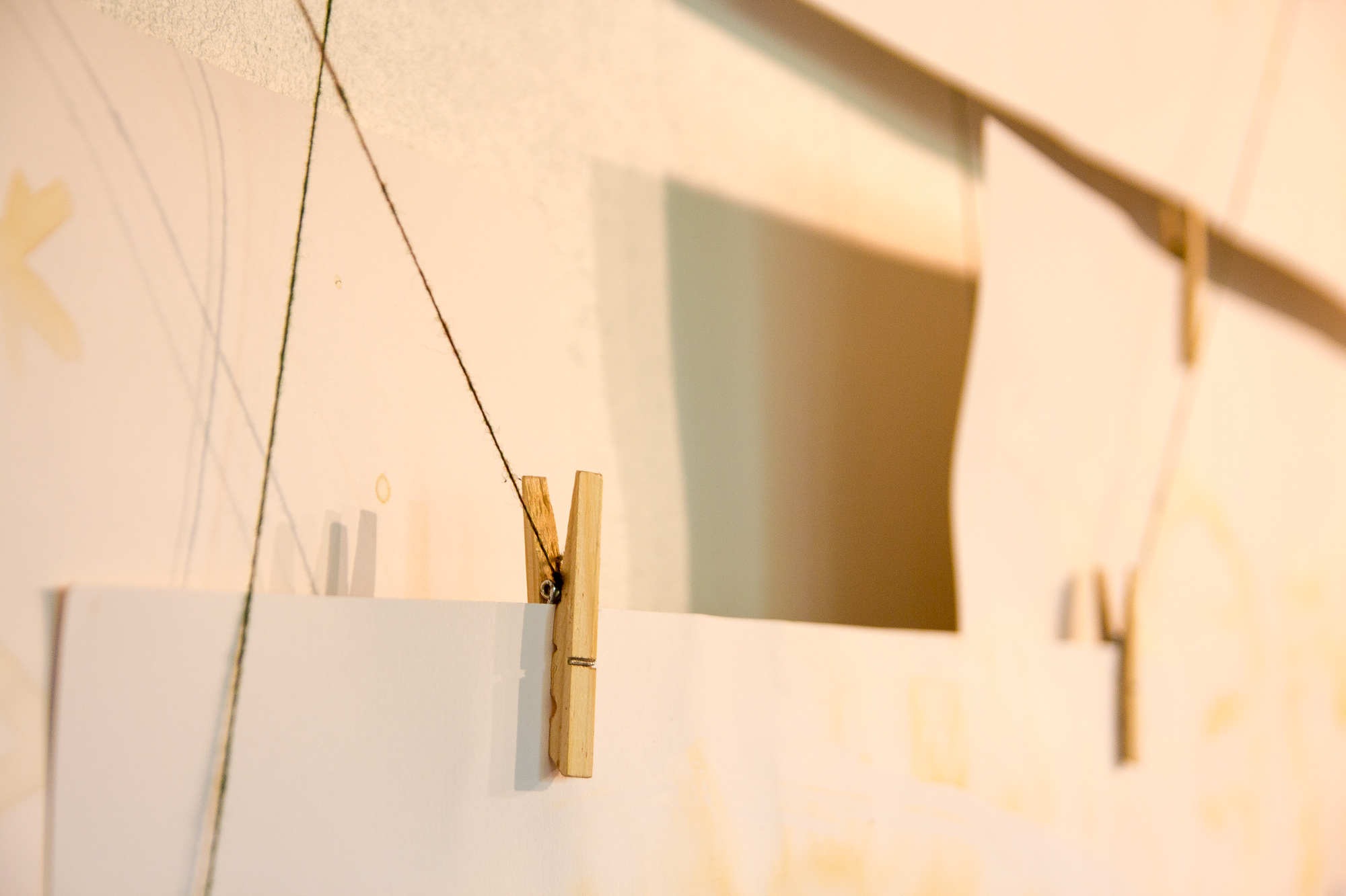 Close-up of two wooden clothespins clipped to a thin string, holding sheets of white papīra. The string is suspended horizontally, casting soft shadows on a beige background. The image is softly lit, highlighting the textures of the clothespins and the papīra.