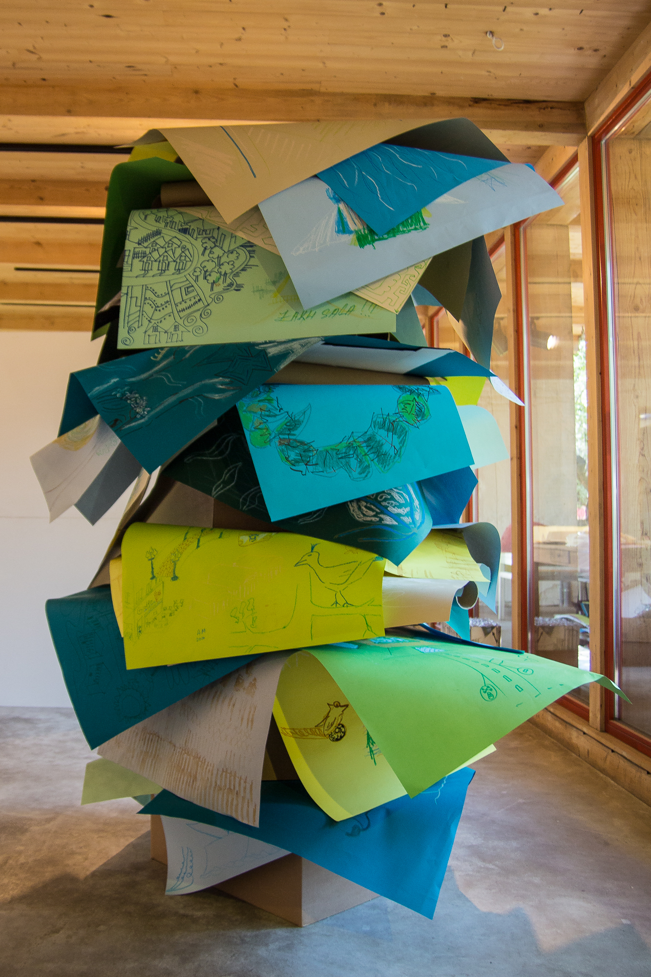 A towering art installation composed of large, colorful sheets of paper, layered and stacked haphazardly. Each sheet features intricate, hand-drawn sketches and diagrams. The structure sits in a wooden-floored gallery with large windows, allowing natural light to illuminate the display at the Papīra Objektu Festivāls.