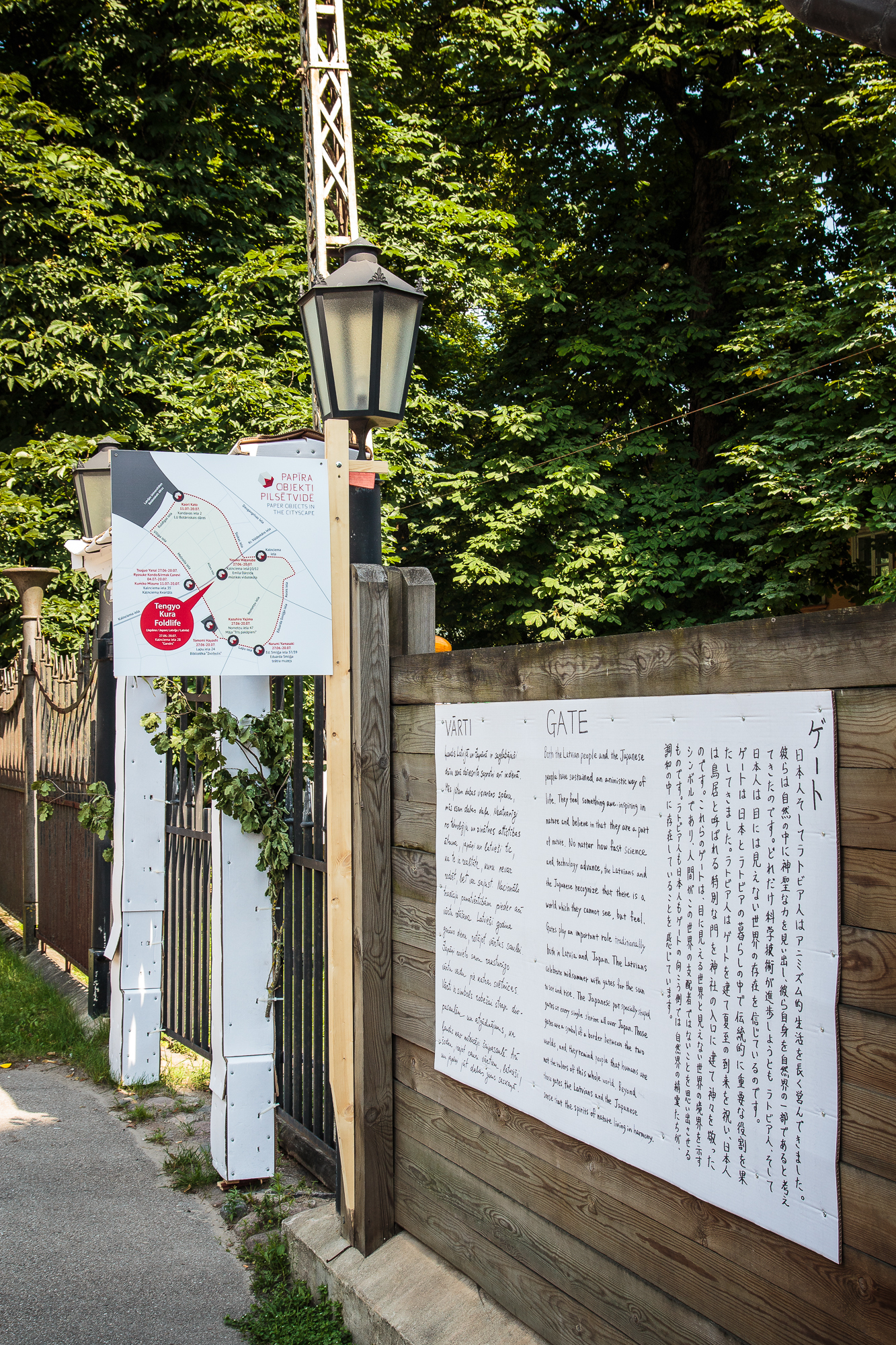 A pathway surrounded by green trees leads to a signboard displaying a map with several markings. To the right of the pathway, a wooden fence supports a white board featuring text in multiple languages about an upcoming festivāls. A vintage lamp post is positioned between the map and the text board.