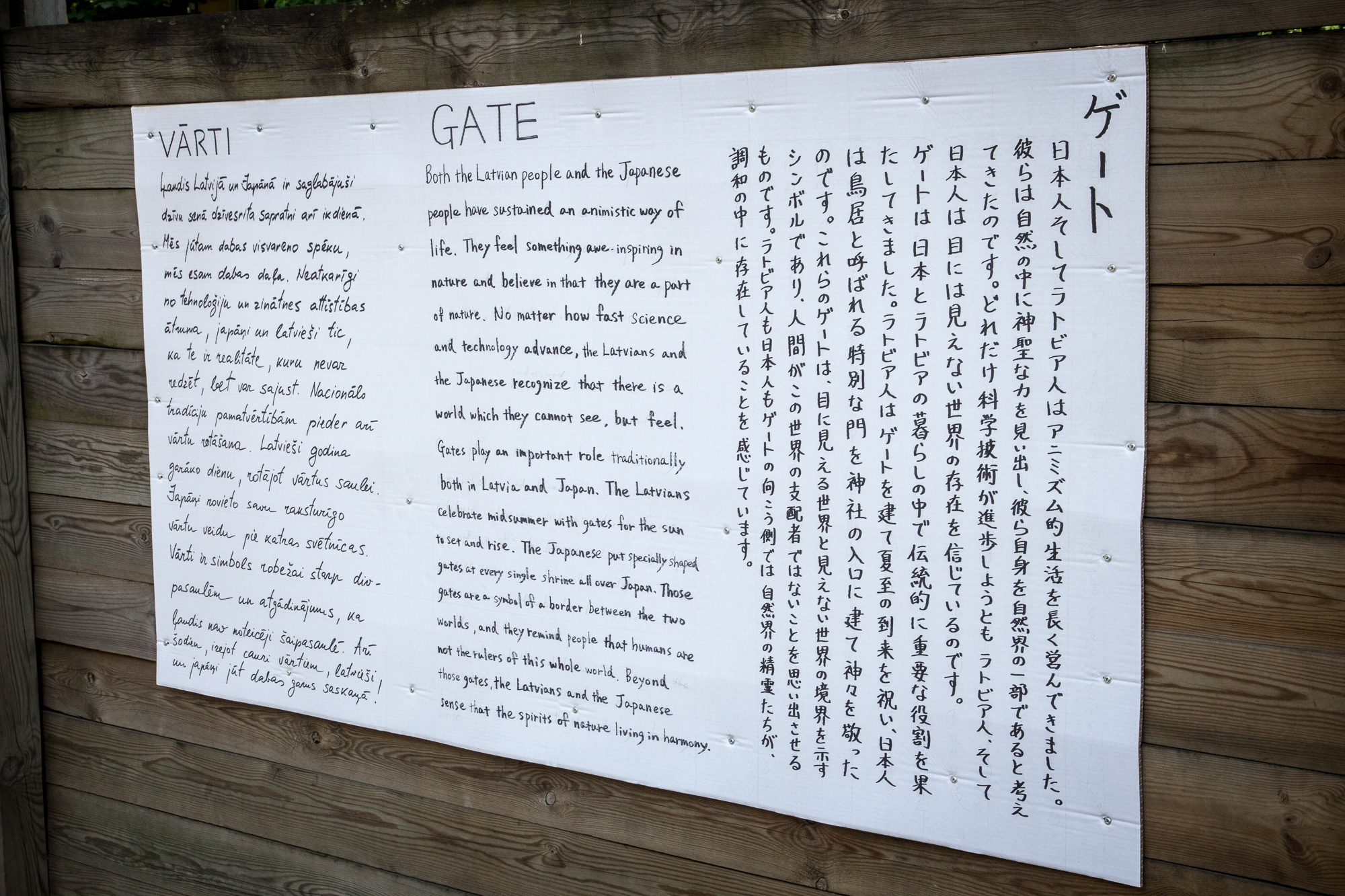 A bilingual exhibition panel featuring text in both European and East Asian languages is pinned to a wooden fence. The white panel with black text meticulously covers historical or informational content about "Gates" and Papīra objektu festivāls in the respective languages.