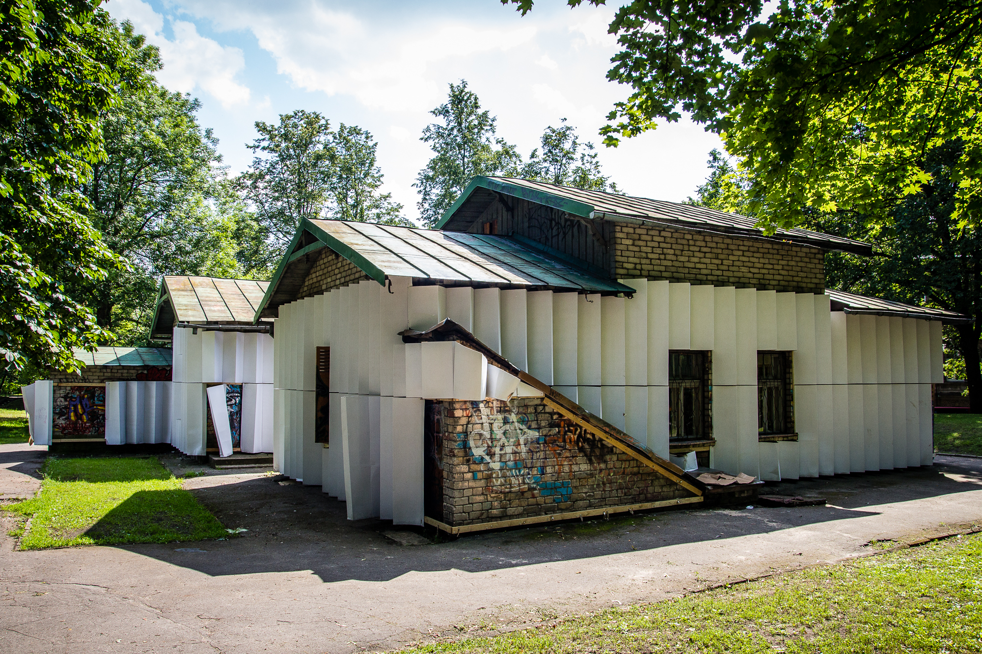 A small brick building with a metal roof and covered in white panels is situated in a park. The structure features several windows and an entrance. Surrounding the building are tall trees and grassy areas, hinting at the creativity of papīra māksla. A graffiti-covered wall is partially visible on one side. The sky is clear and sunny.