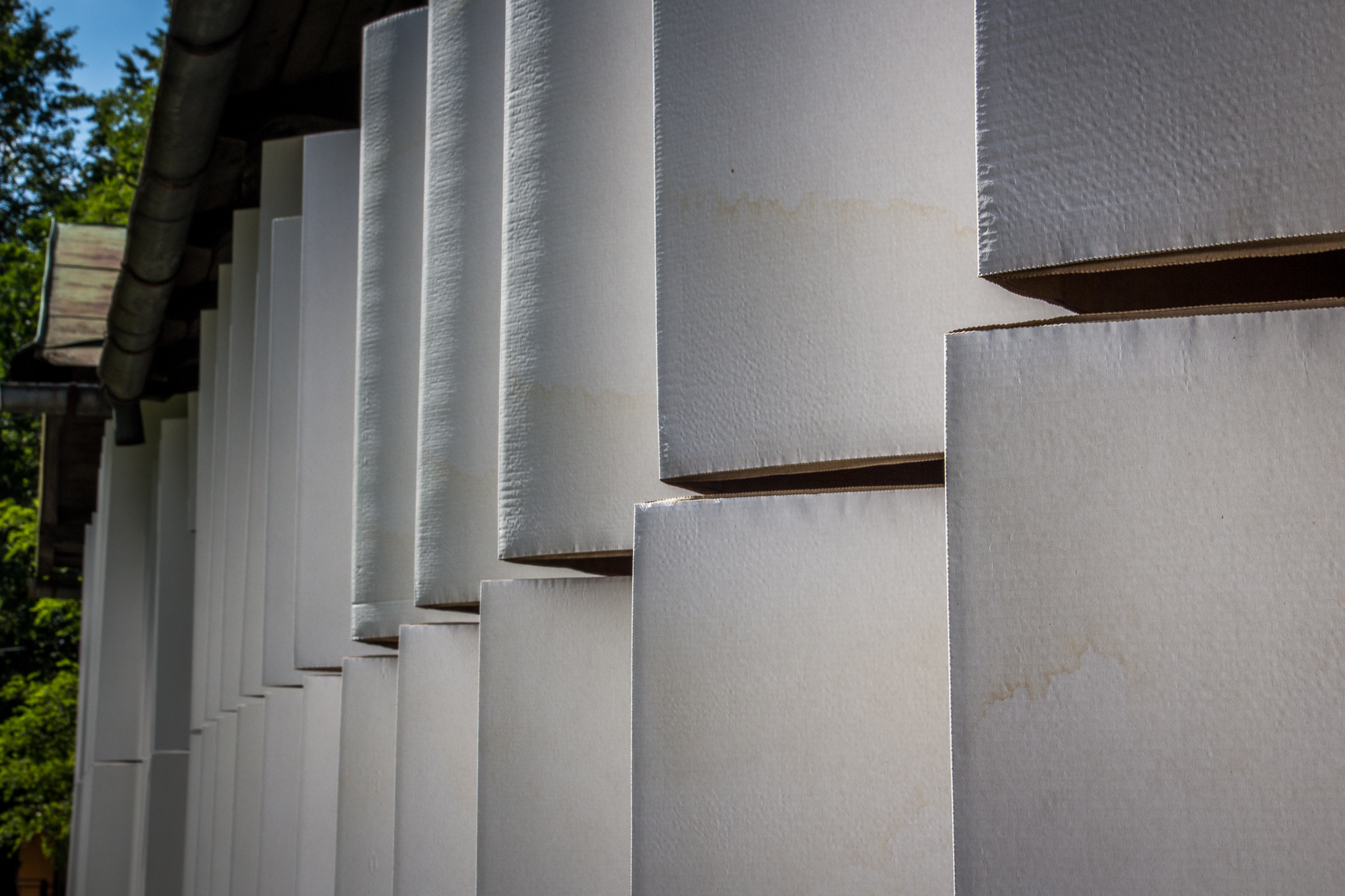 A close-up view of an architectural feature with a series of vertical, rectangular white panels arranged in a staggered pattern reminiscent of traditional Papīra artwork. The panels show signs of weathering with visible stains and marks. Part of a roof with a gutter is visible on the left against a background of green foliage.
