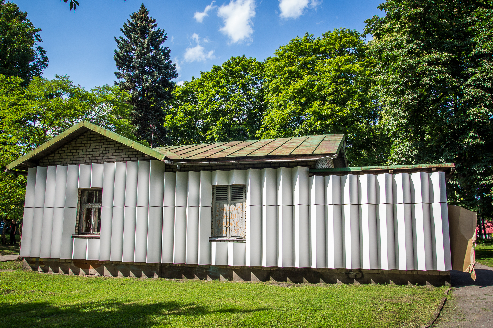 A small house with a corrugated metal exterior stands in a grassy area surrounded by tall, leafy trees. The roof appears weathered, showing signs of rust. Bright sunlight casts shadows on the house and the green grass. A path runs beside the house, reminiscent of routes taken during the Papīra objektu festivāls.