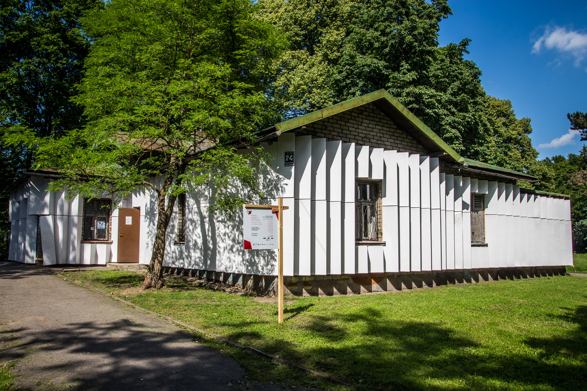 A white building with vertical paneling stands surrounded by greenery. A tree with dense foliage shades the left side, partially obscuring a door. A wooden signpost promoting the Papīra Objektu Festivāls is to the right of the door. The sky is clear and blue, casting sharp shadows on the grass and path in the foreground.