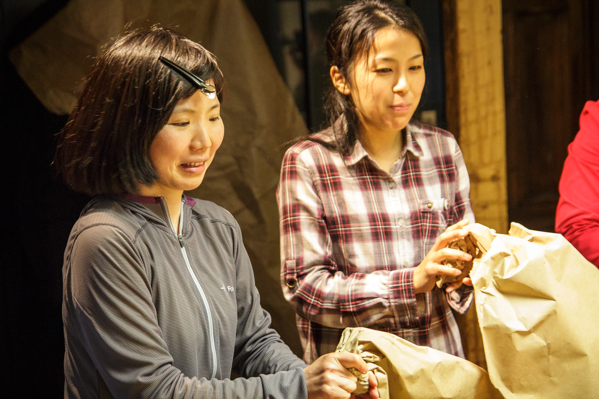 Two women are standing indoors, engaged in a task. The woman on the left is wearing a headlamp and a gray jacket, focused on an objektu in front of her. The woman on the right is in a red and gray plaid shirt, also looking down as she works, holding a brown papīra bag.