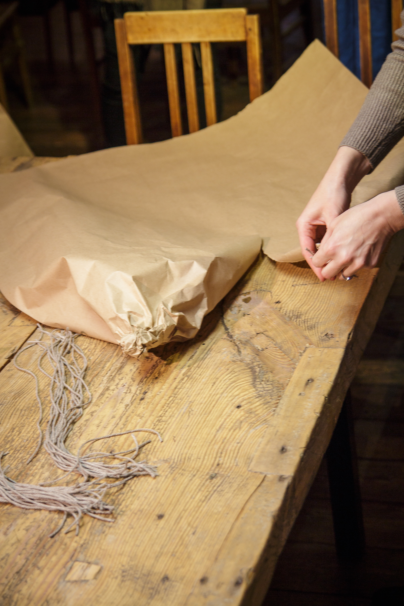 A pair of hands is seen wrapping a Papīra objektu in brown paper on a wooden table. Twine is spread out on the table, ready to be used for securing the package. In the background, there are wooden chairs, indicating a rustic setting. The scene suggests a process of packing or gift wrapping for an upcoming festivāls.