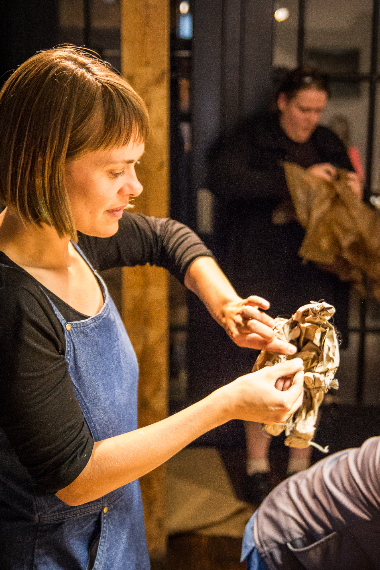 Woman in a denim apron smiling and shaping a piece of brown paper in her hands, with another person in the background also handling brown paper. They appear to be engaged in a Papīra craft activity, possibly preparing for an objektu festivāls, in a warmly lit indoor setting.