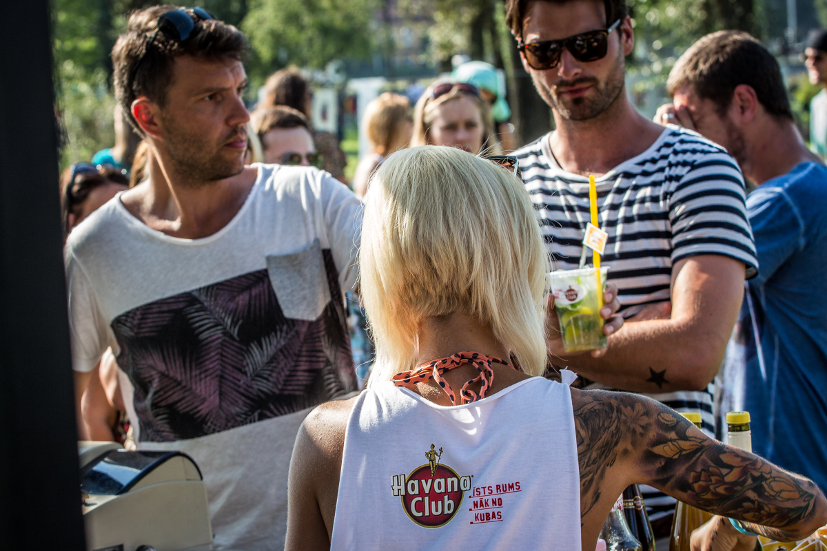 A crowd of people enjoying an outdoor event. At the foreground, a woman with platinum blonde hair and tattoos, wearing a white "Havana Club" top, is serving drinks. The man in front of her is speaking while holding a drink. Another man in sunglasses and a striped shirt stands nearby at Piens Fest 2014.