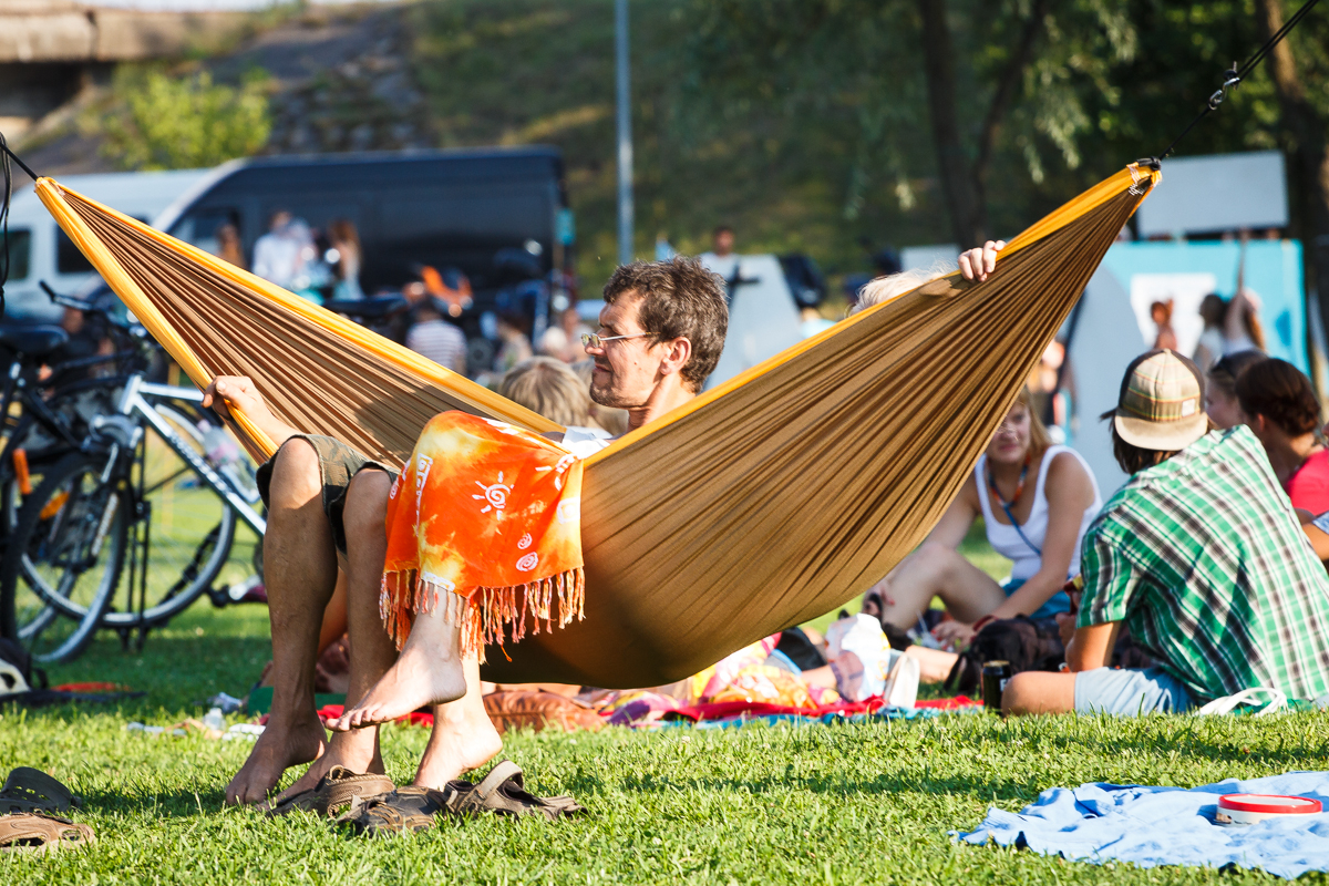 A man is lying in a brown hammock, reading a book at Piens Fest 2014, with an orange and yellow patterned blanket over his legs. People are sitting on the grass around him, some on blankets. Bicycles and a black vehicle are in the background. It's a sunny day with green grass and trees visible.