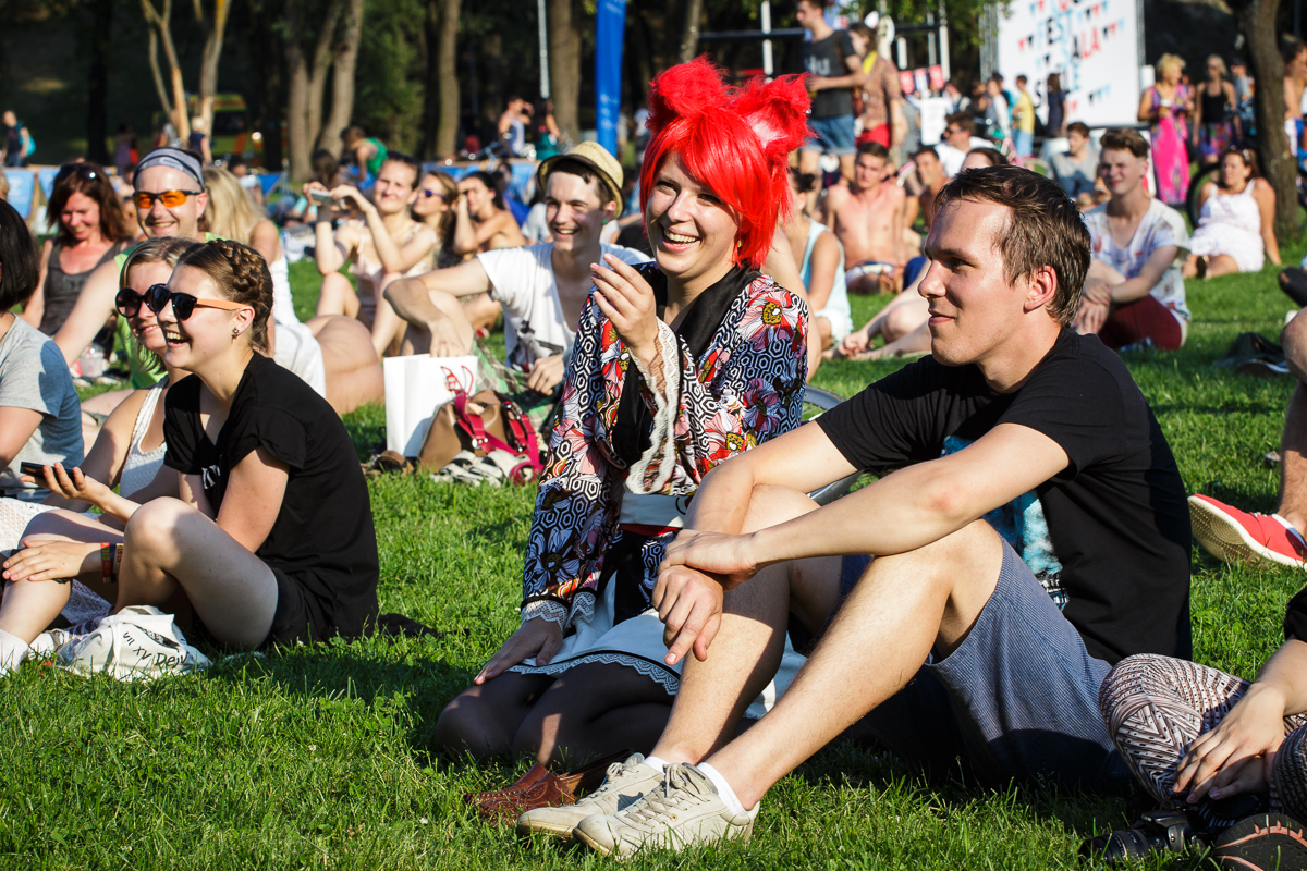 At PiensFest 2014, a group of people is sitting on the grass at an outdoor event on a sunny day. They appear to be enjoying the fest, with some smiling and laughing. One person in the center stands out with bright red hair and colorful clothing. Trees and other attendees are visible in the background.