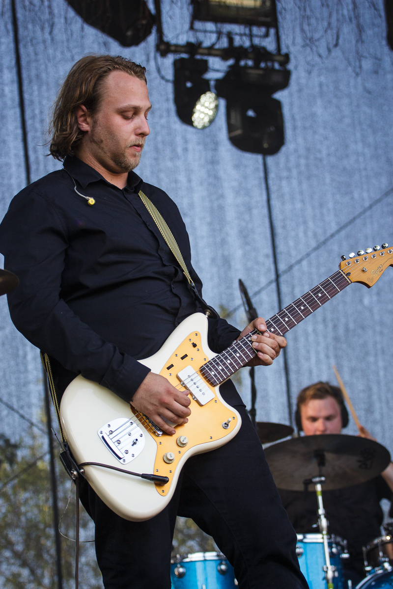 A musician with shoulder-length hair plays an electric guitar on stage at Piens Fest 2014. He wears a black shirt and has a strap over his shoulder. In the background, another musician is visible playing drums. Stage lights and draped netting create ambiance in the setting.