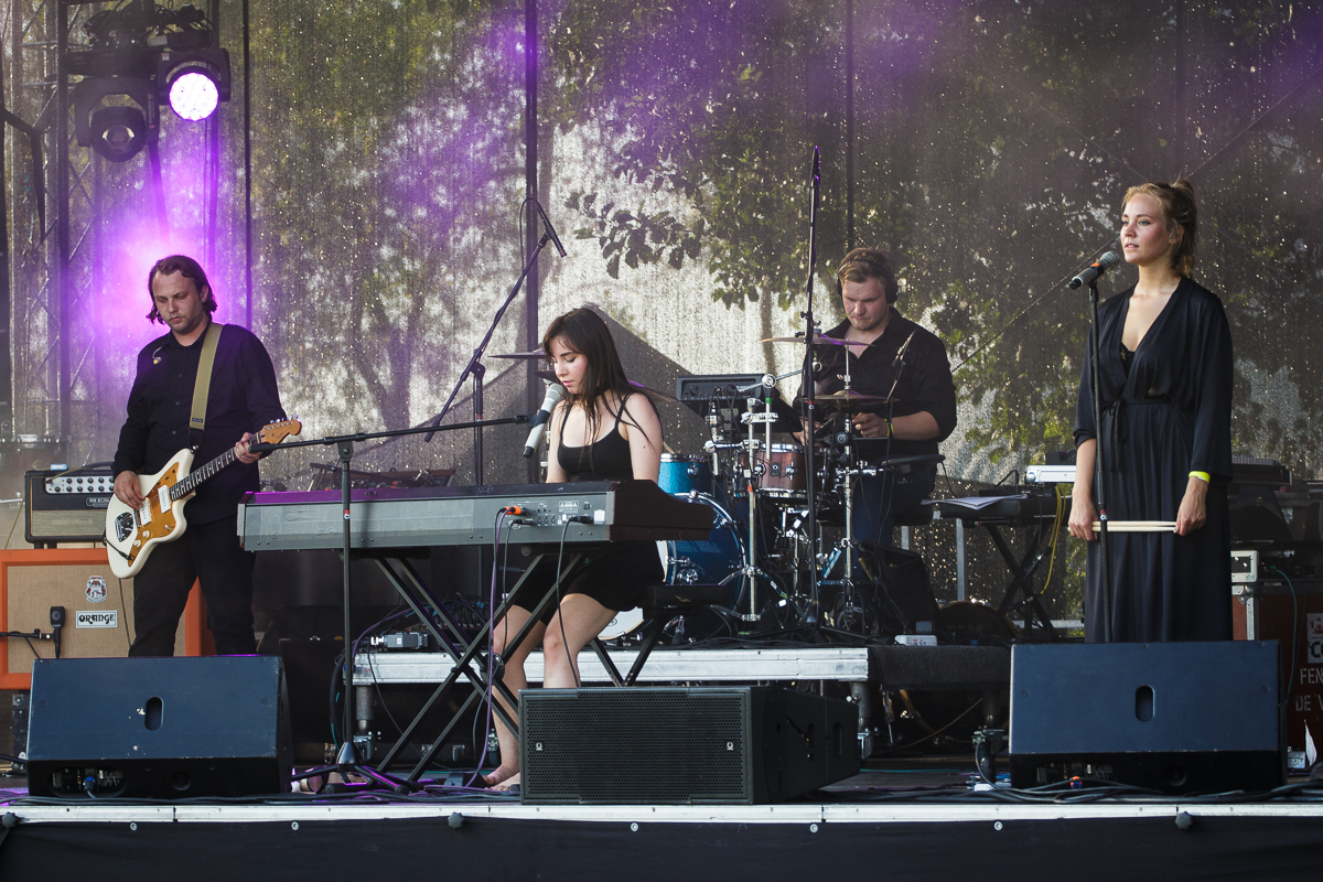 A band performs on an outdoor stage at Piens Fest. The group includes a keyboardist, a bassist, a drummer, and a female vocalist. The keyboardist and vocalist are at the front, with the drummer behind and the bassist to the left. Purple stage lights illuminate the scene, and trees are visible in the background.