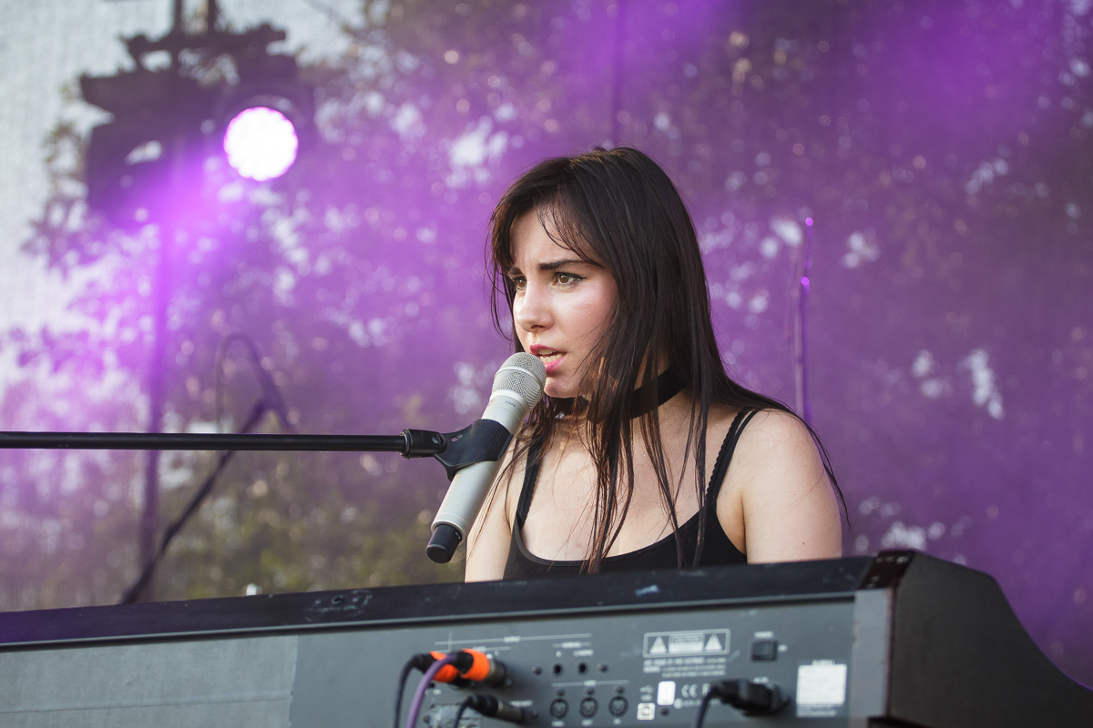 A woman with long dark hair, wearing a sleeveless black top, is singing into a white microphone while playing a keyboard on an outdoor stage at the 2014 Piens Fest. The background features purple lighting and blurred foliage. She appears focused and immersed in her performance.
