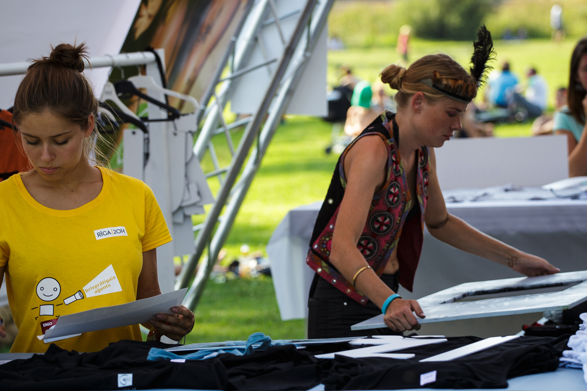 At Piens Fest 2014, two women at an outdoor event work at separate tables with various materials. The woman on the left, wearing a yellow shirt and name tag, appears to be organizing or inspecting items. The woman on the right, in a colorful vest and headband with a feather, arranges materials on her table.