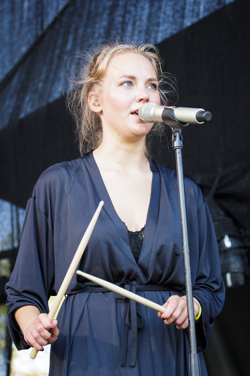 A woman with blonde hair tied back is singing into a microphone on a stand. At Piens Fest 2014, she is holding two drumsticks and wearing a dark, loose-fitting outfit with a tied waist. The background is a black stage setup. The image captures her in a performance setting.